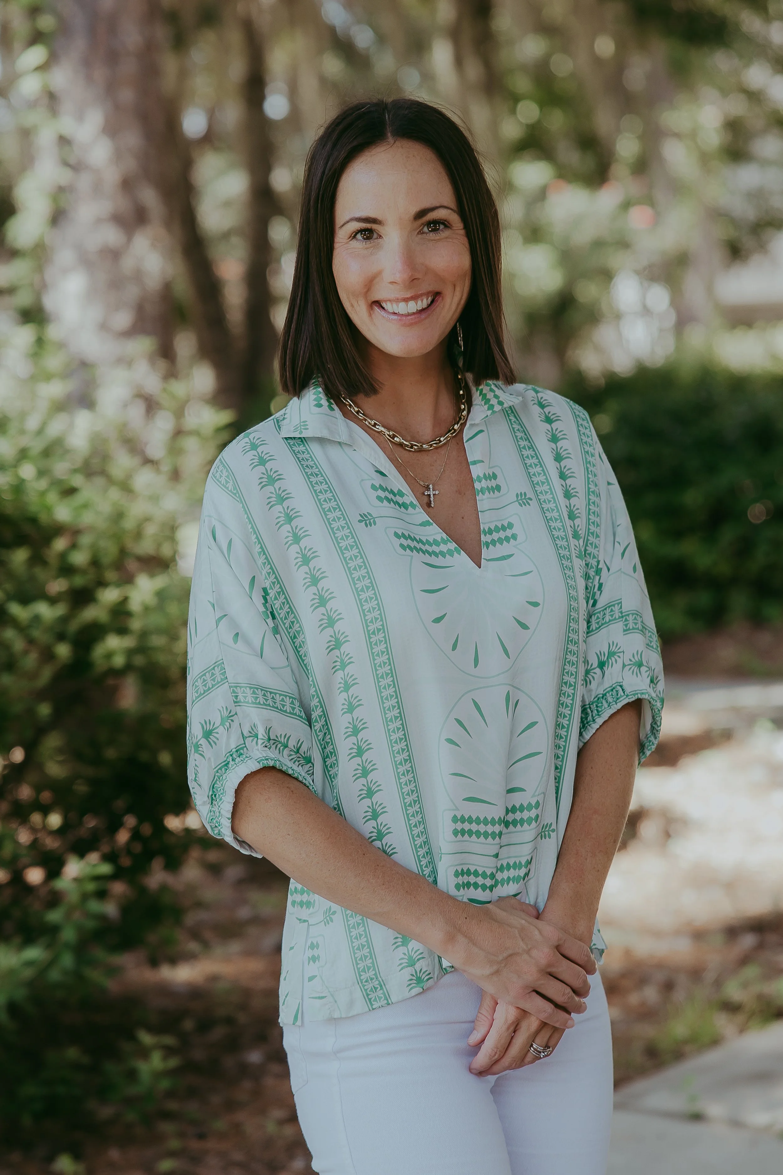 A woman with shoulder-length dark hair smiling outdoors, wearing a white and green patterned blouse with white pants, accessorized with layered necklaces, including a cross, and standing in a garden with trees and bushes in the background.