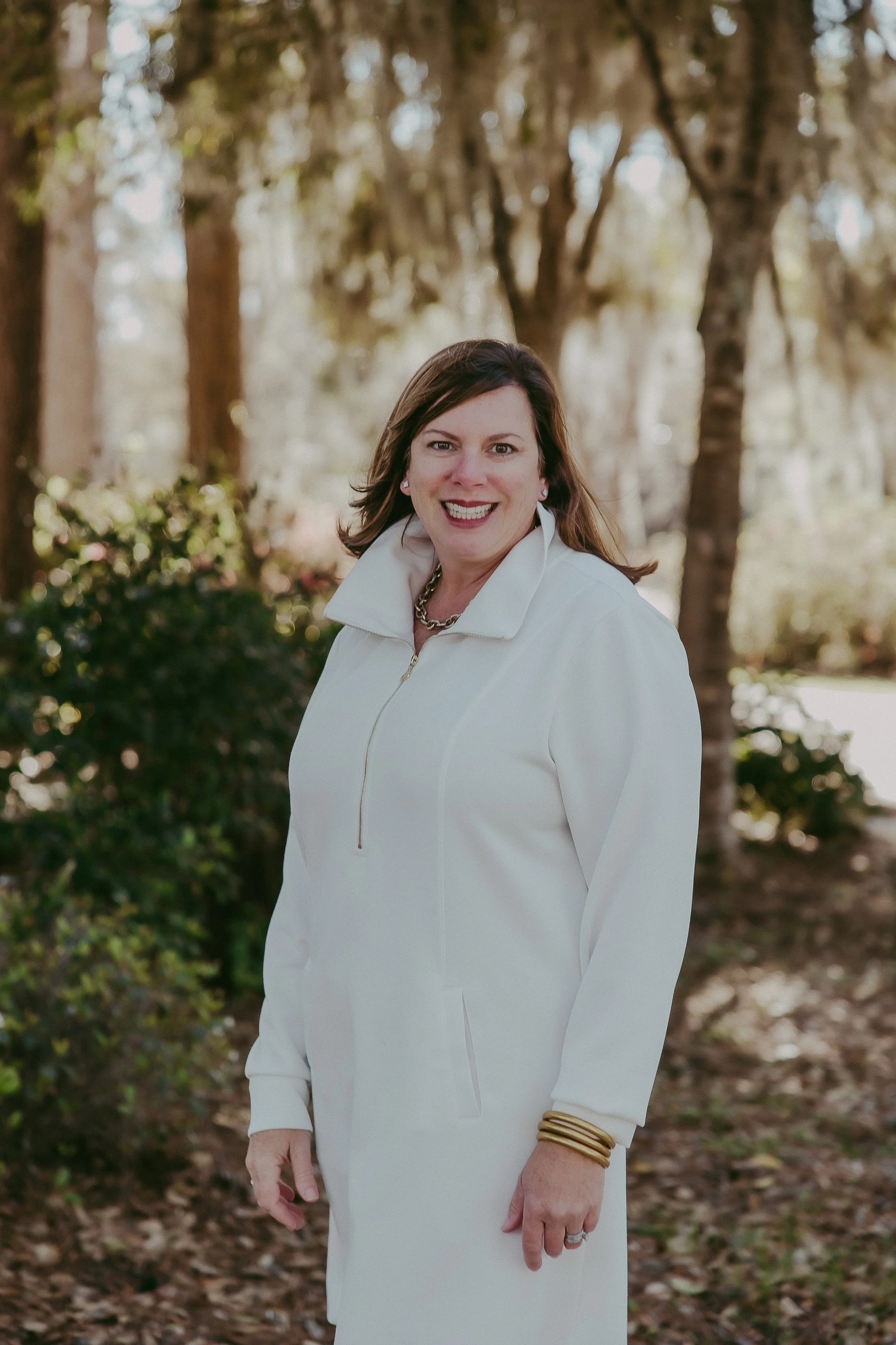A woman smiling outdoors in front of trees and greenery, wearing a white coat and gold jewelry.