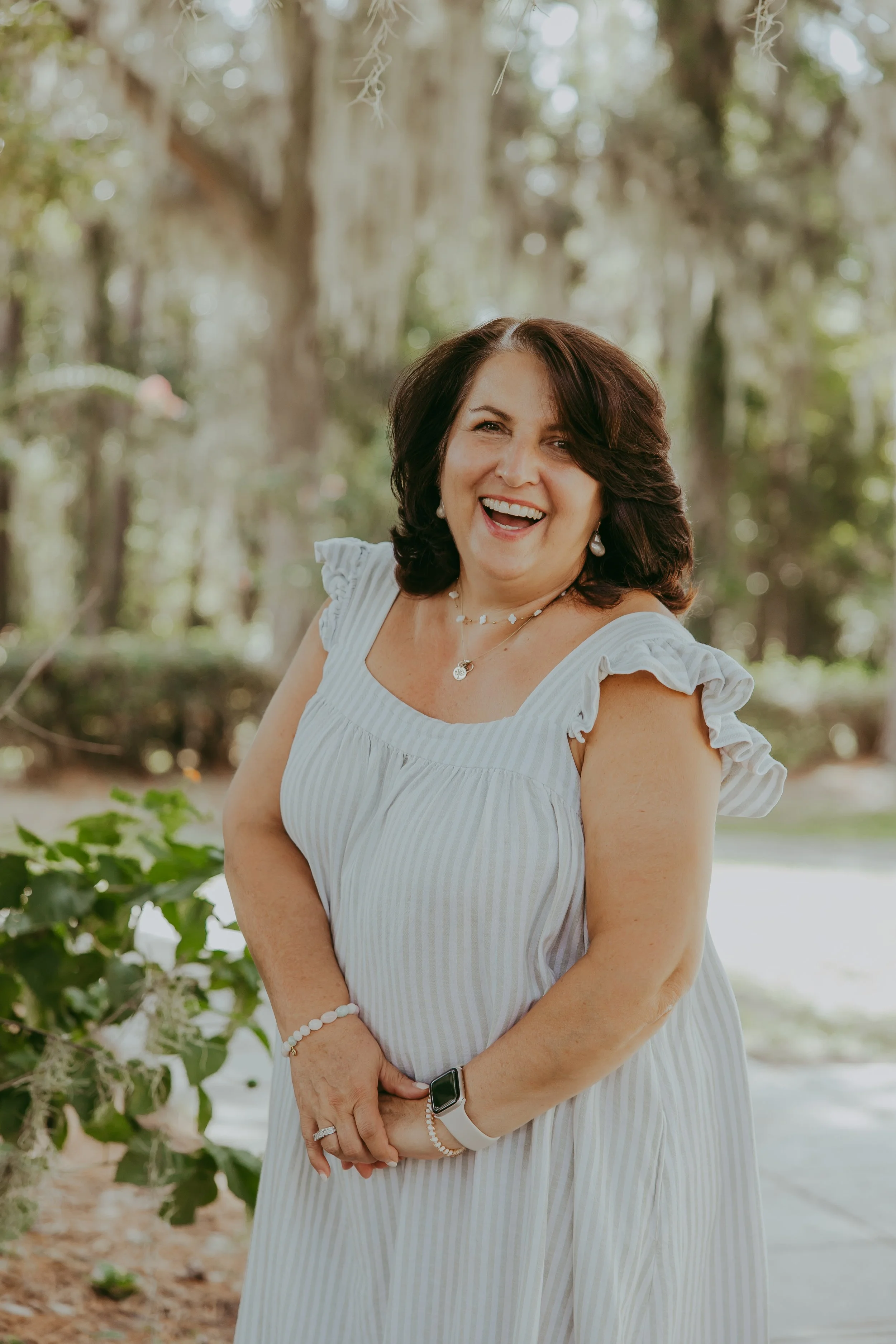 A woman with dark hair smiling outdoors, wearing a navy blue blouse with white trim and white pants.