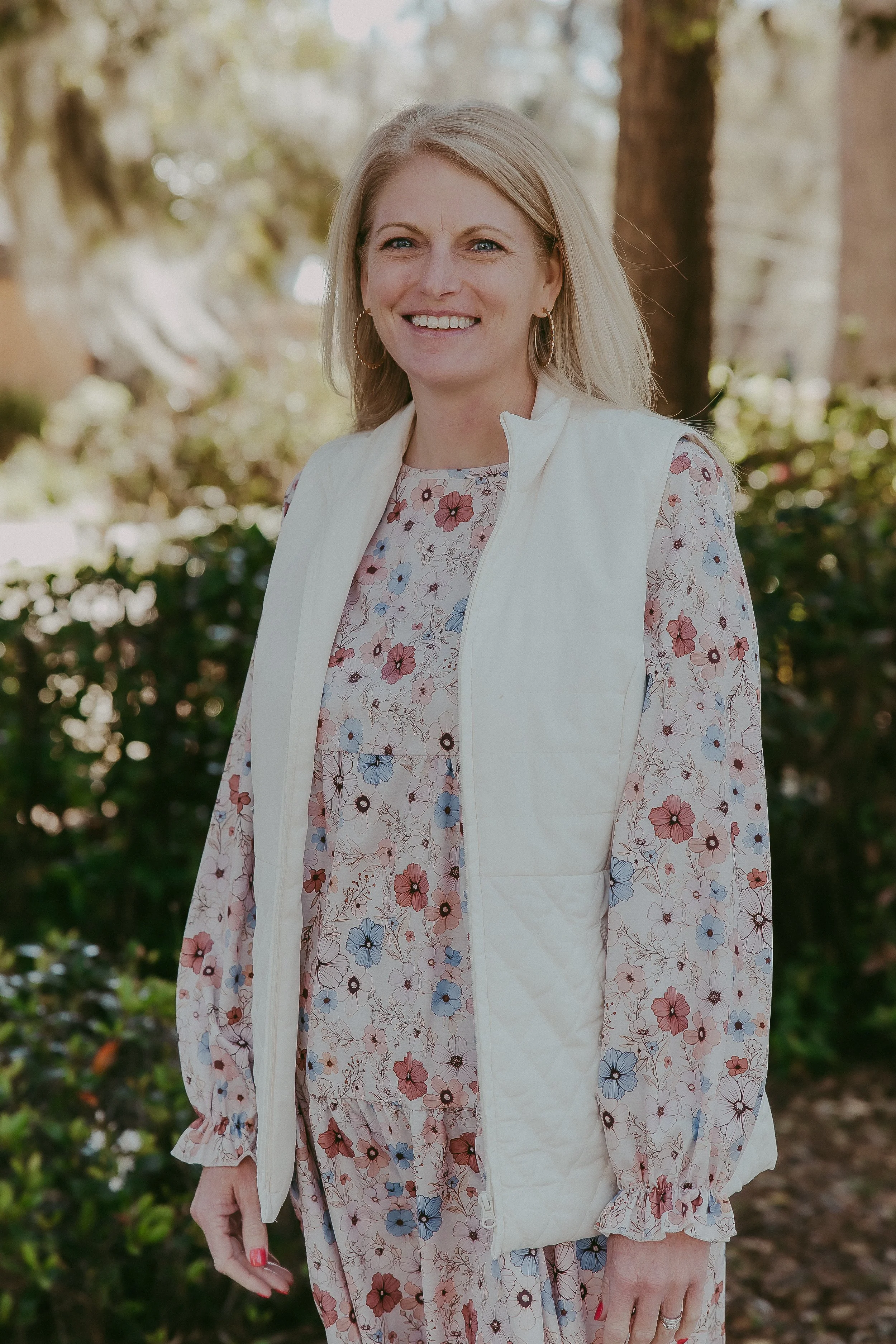 A woman with blonde hair in a floral dress and a white vest standing outdoors with trees and greenery in the background.