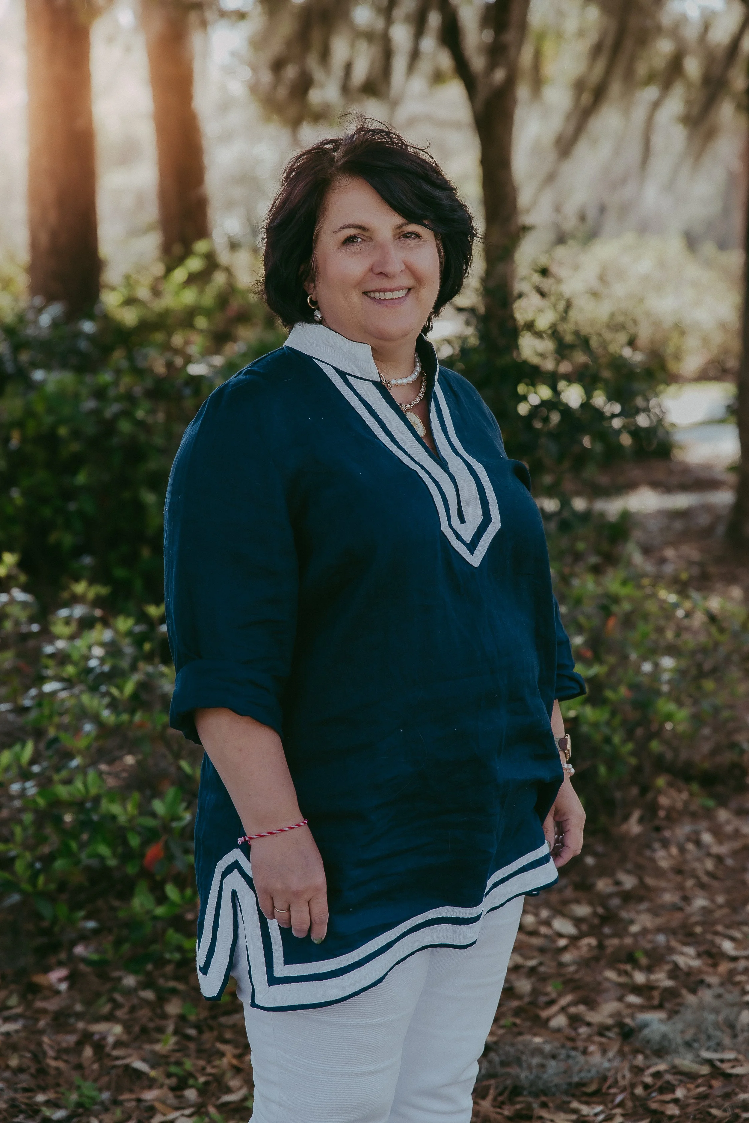 A woman with dark hair smiling outdoors, wearing a navy blue blouse with white trim and white pants.
