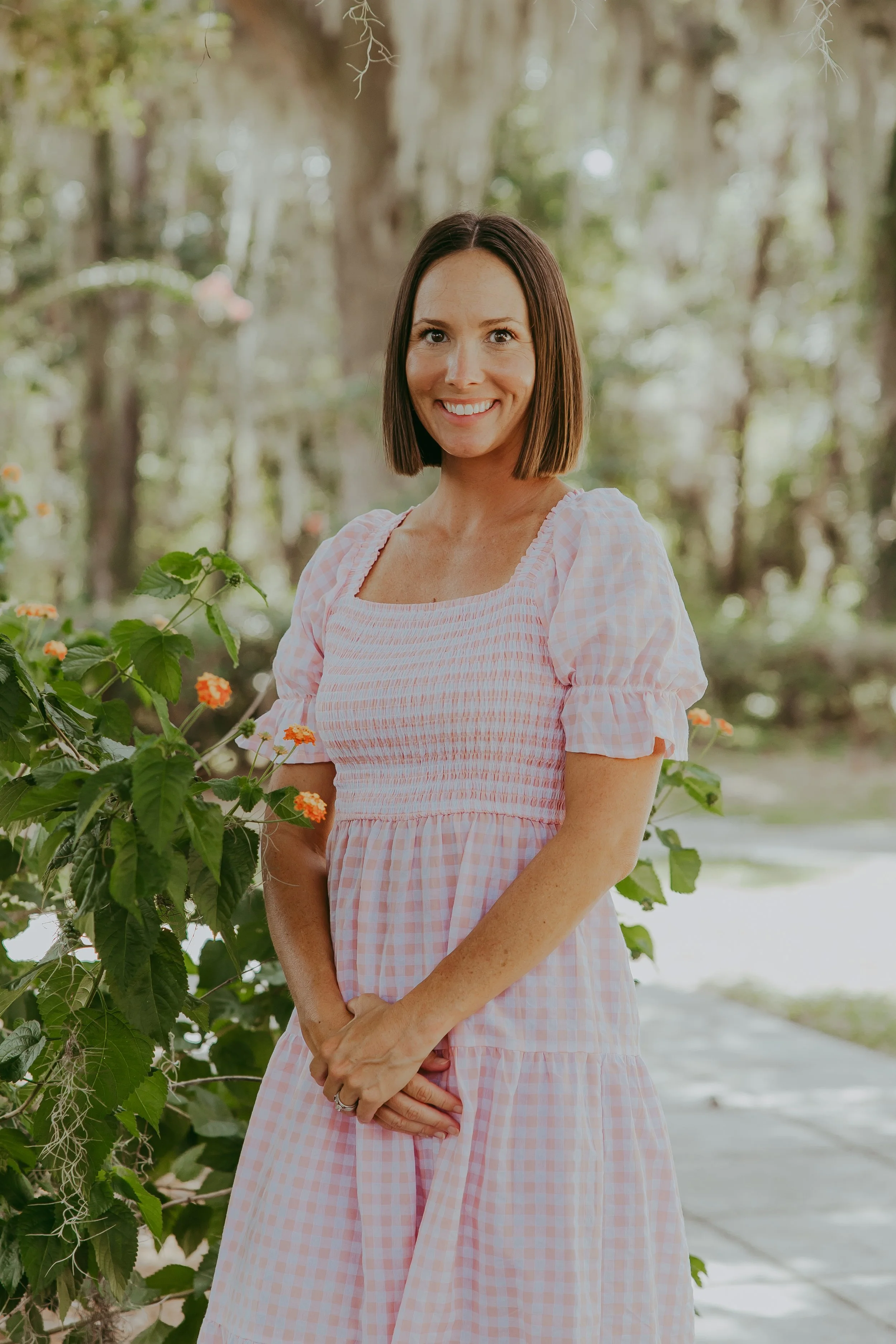 A woman with shoulder-length dark hair smiling outdoors, wearing a white and green patterned blouse with white pants, accessorized with layered necklaces, including a cross, and standing in a garden with trees and bushes in the background.