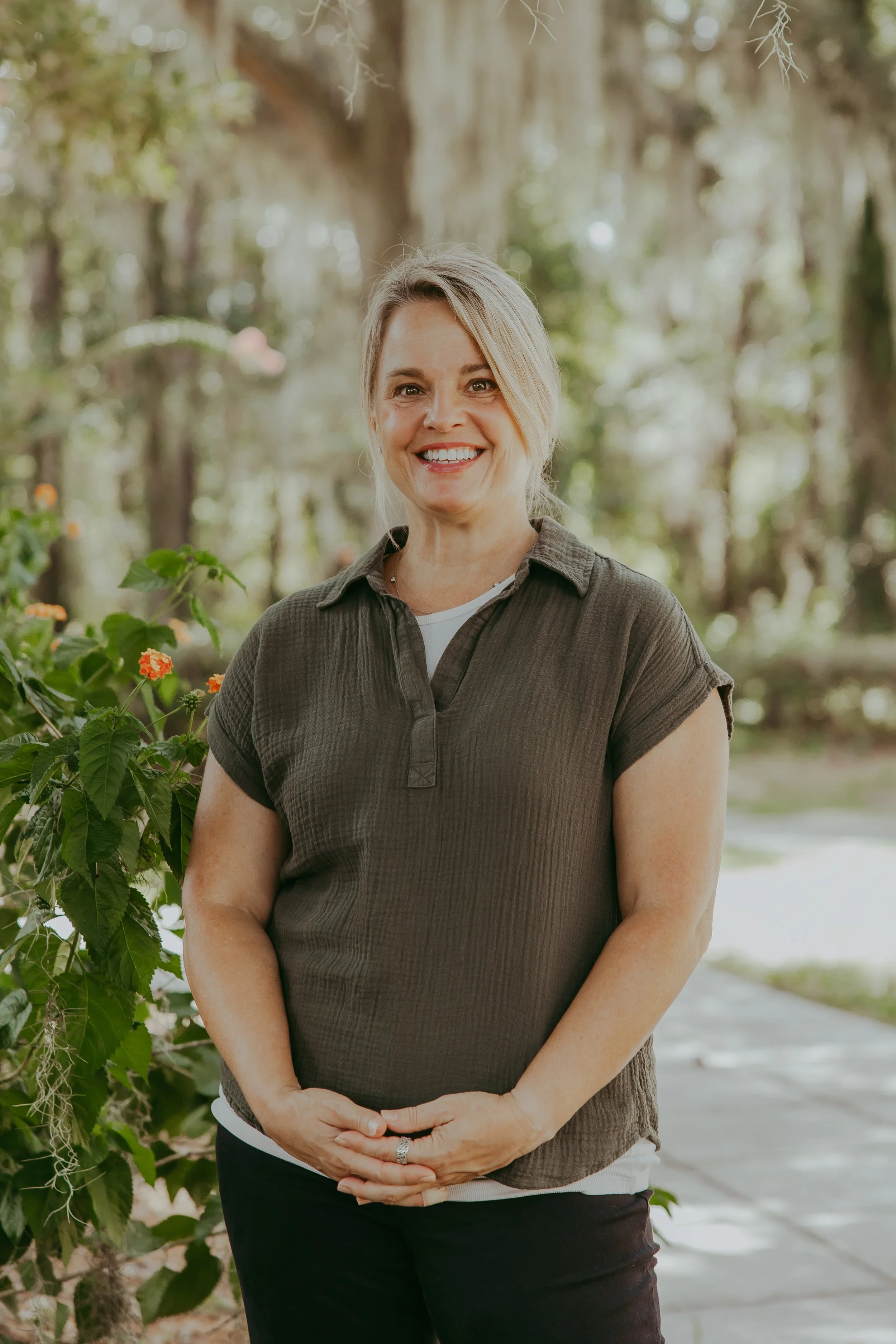 A woman with light skin and shoulder-length brown hair, smiling and wearing a sleeveless pink top and dark shorts, standing outdoors with green foliage and trees in the background.