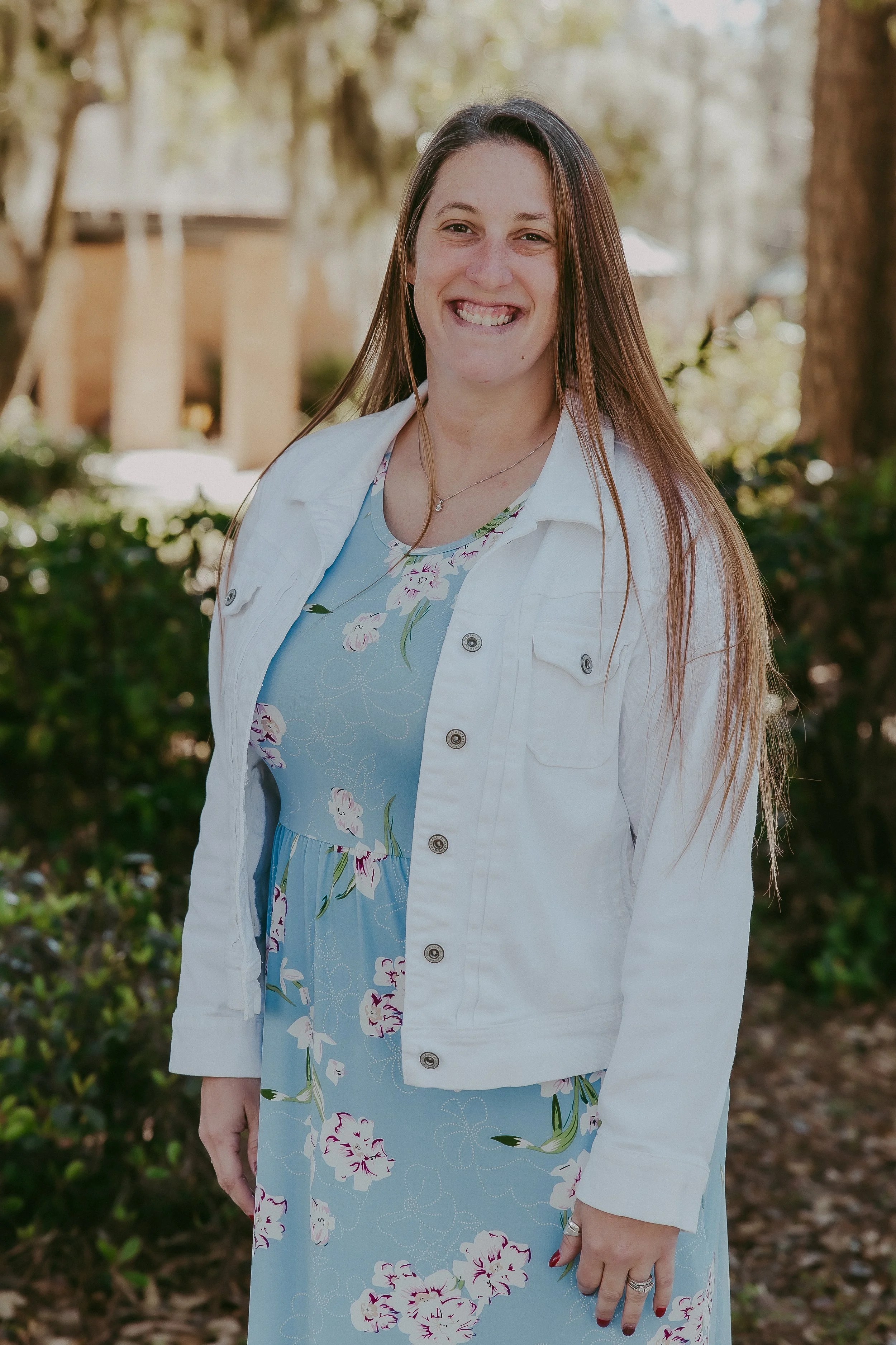 A woman smiling outdoors, wearing a white denim jacket over a light blue floral dress, standing in front of trees and greenery.