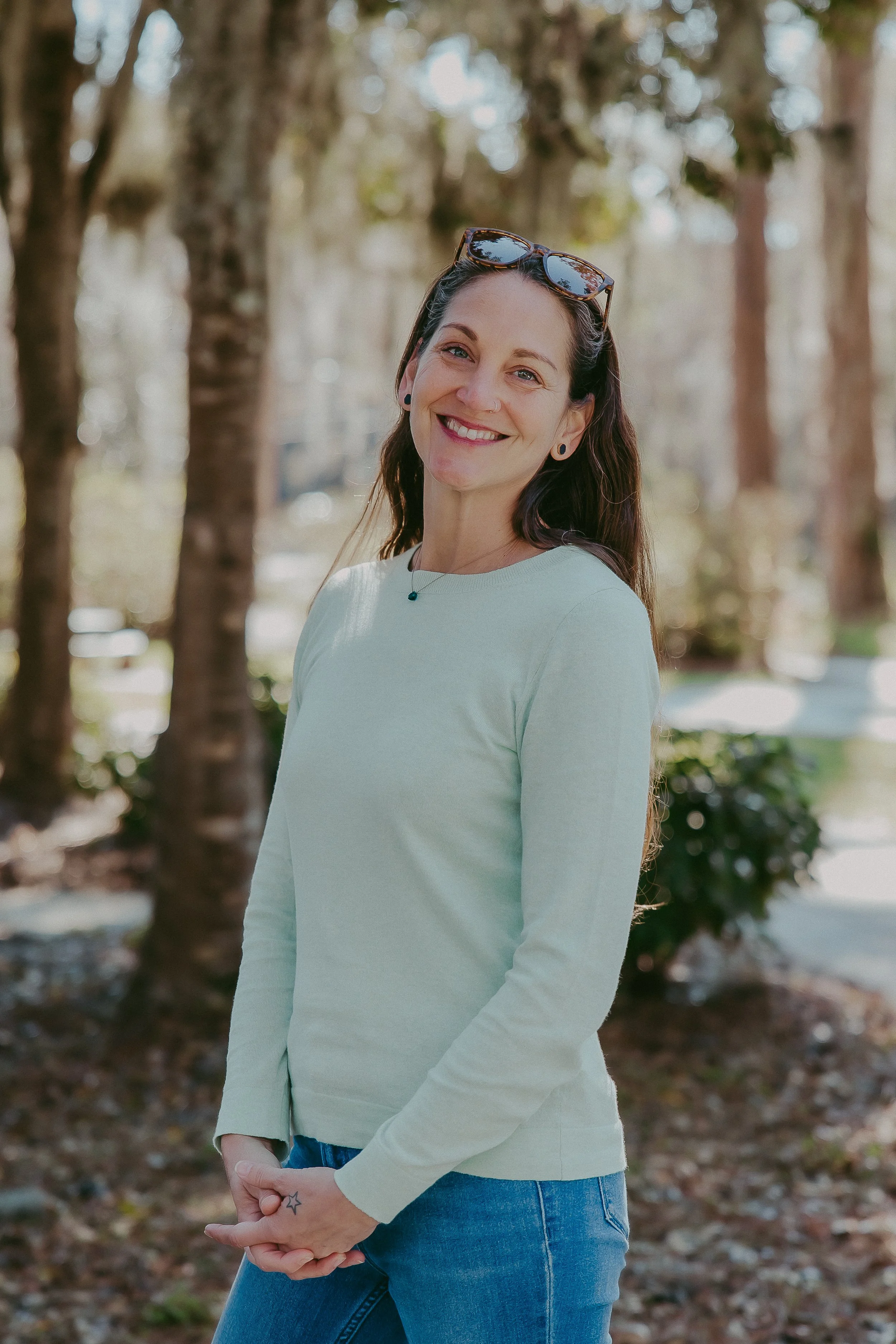 A woman smiling outdoors in a park with trees in the background, wearing sunglasses on her head, a light-colored long-sleeve shirt, and blue jeans.