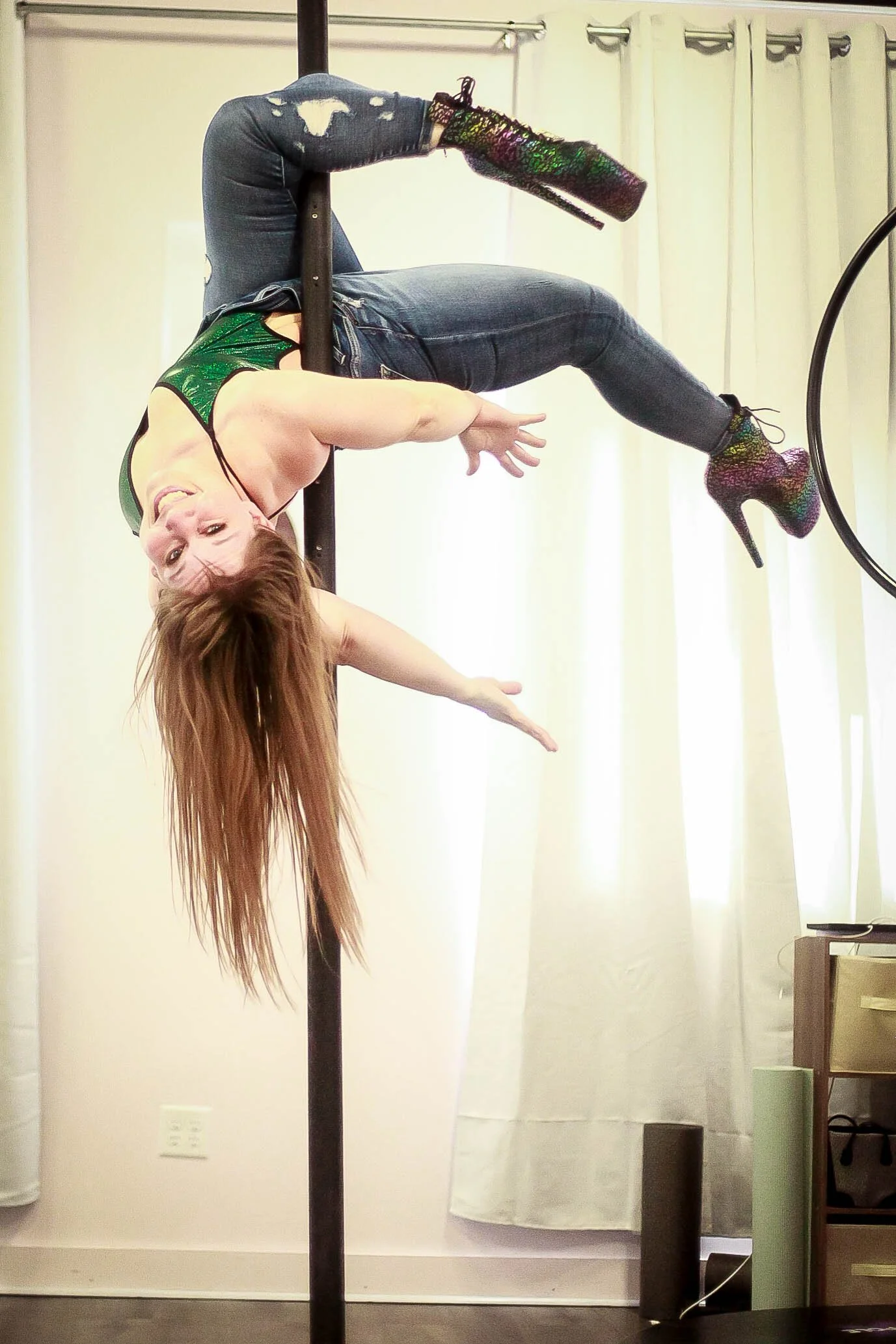 Woman with long hair performing a pose on a dance pole, wearing a green top, ripped jeans, and sparkly high-heeled boots, indoors with curtains and furniture in the background.