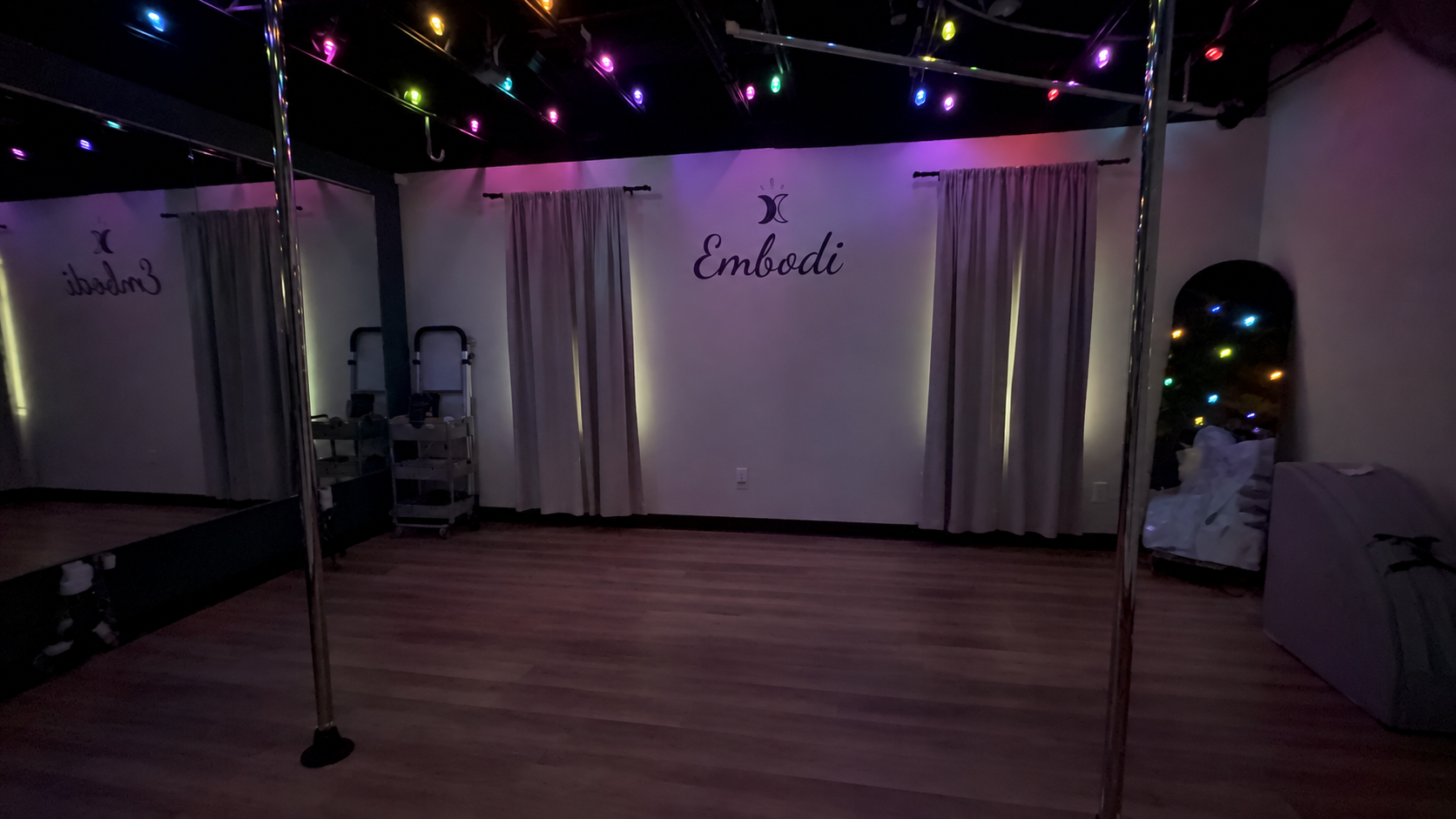 Dark, intimate pole fitness studio at Embodi Wellness Studio in downtown Hudson, Wisconsin, with three poles, mirrors, wood floors, and colorful ceiling lights.