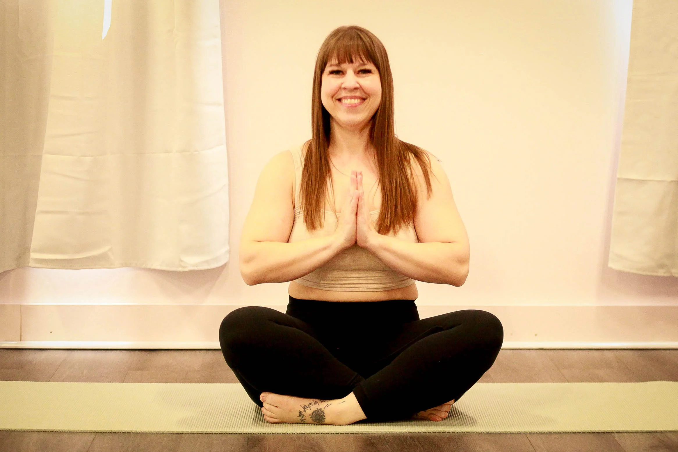 Woman sitting cross-legged on yoga mat, hands in prayer position, smiling, in a yoga studio with curtains.