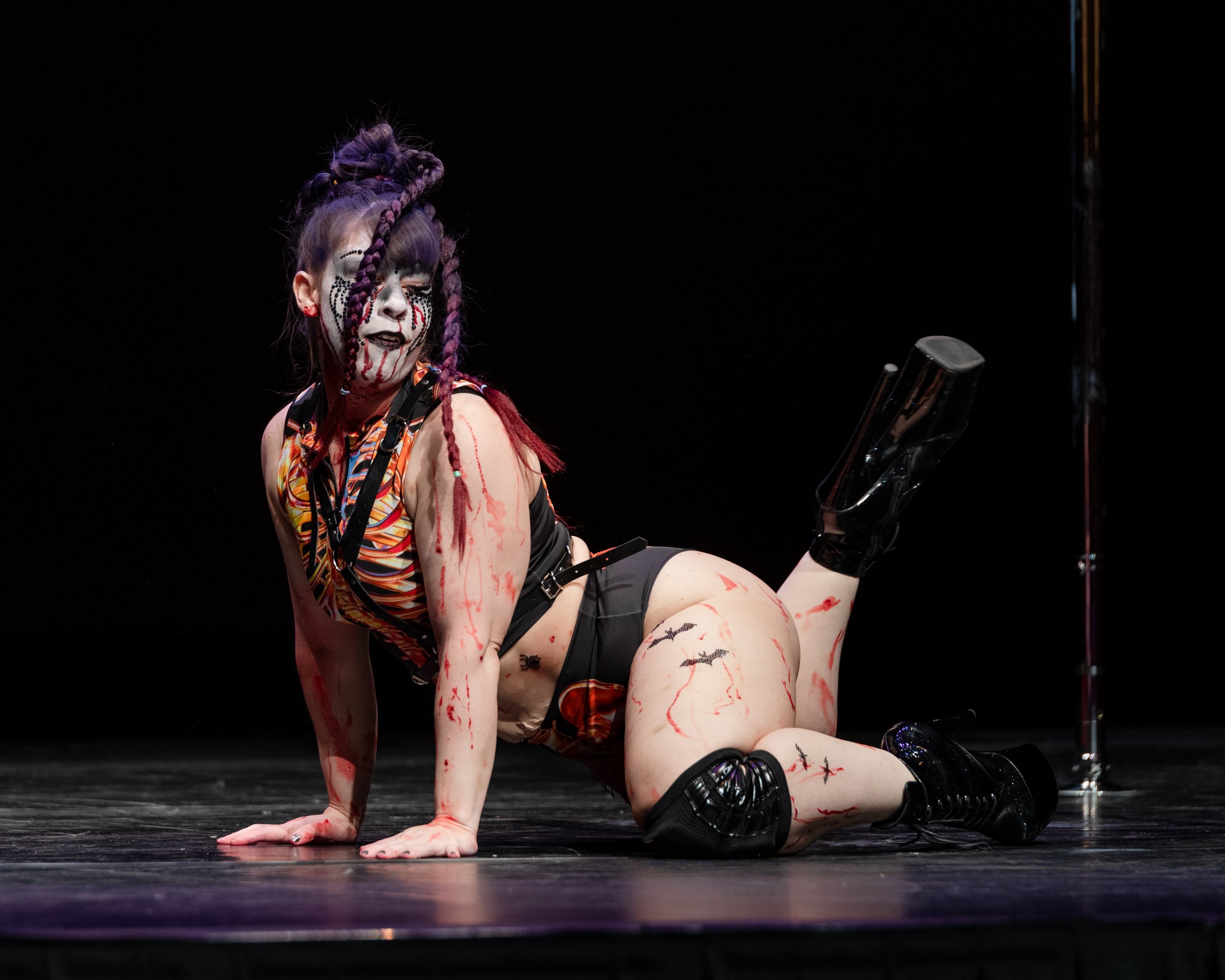 Performer in theatrical costume with elaborate makeup, purple braided hair, and blood-like paint on her skin, crouching on stage with one knee on the ground and one leg bent, wearing combat boots and a colorful top, against a black backdrop.