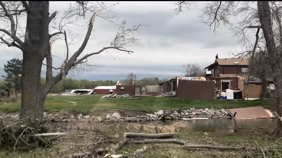 Damaged house and trees near a water body, with a cloudy sky in the background
