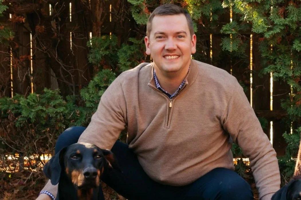 Tom in a tan sweater sitting outdoors with a black and tan dog, a green shrub, and a wooden fence in the background.