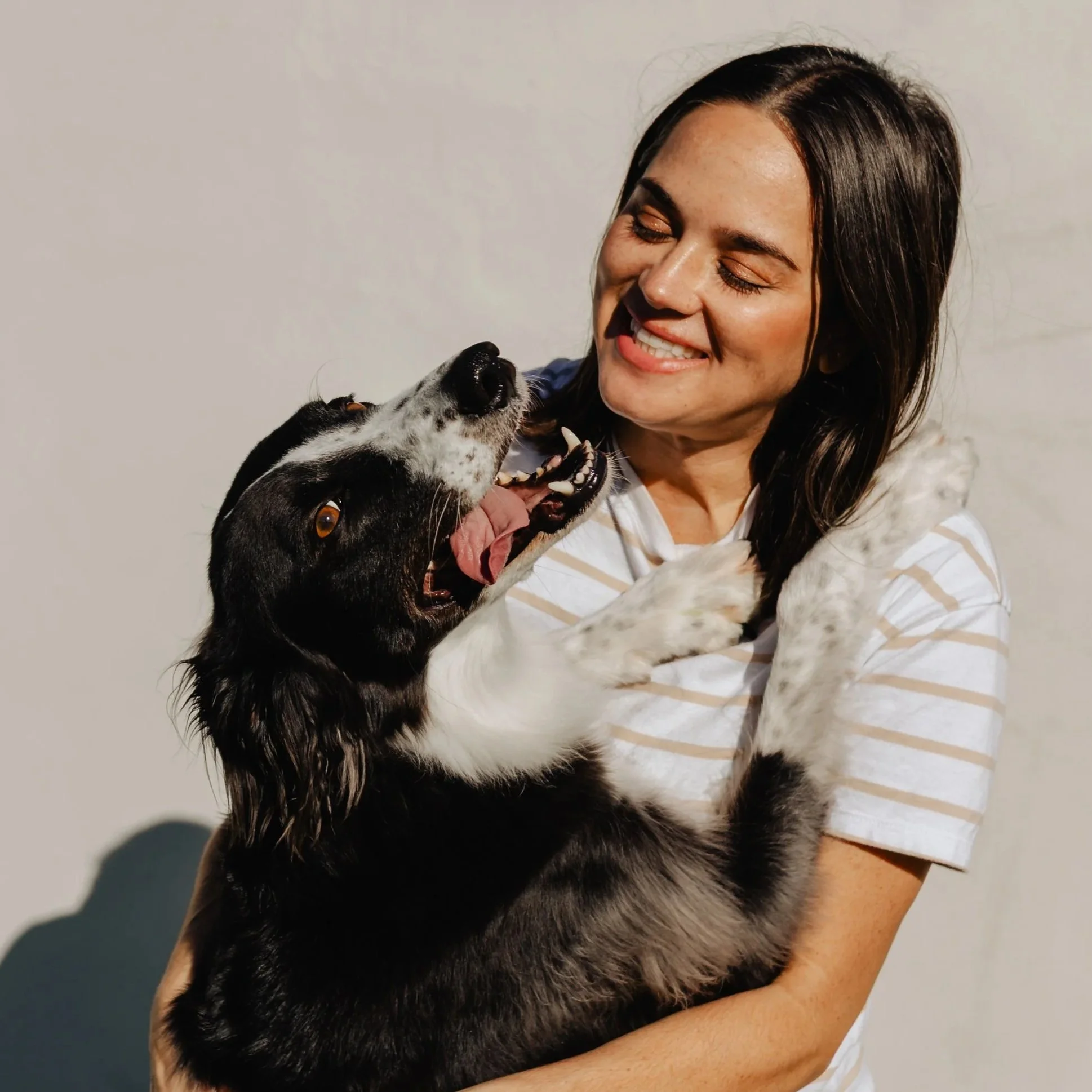 A woman smiling and holding a black and white dog in her arms. The dog is looking up at the woman with its mouth open, tongue out, and appears happy.