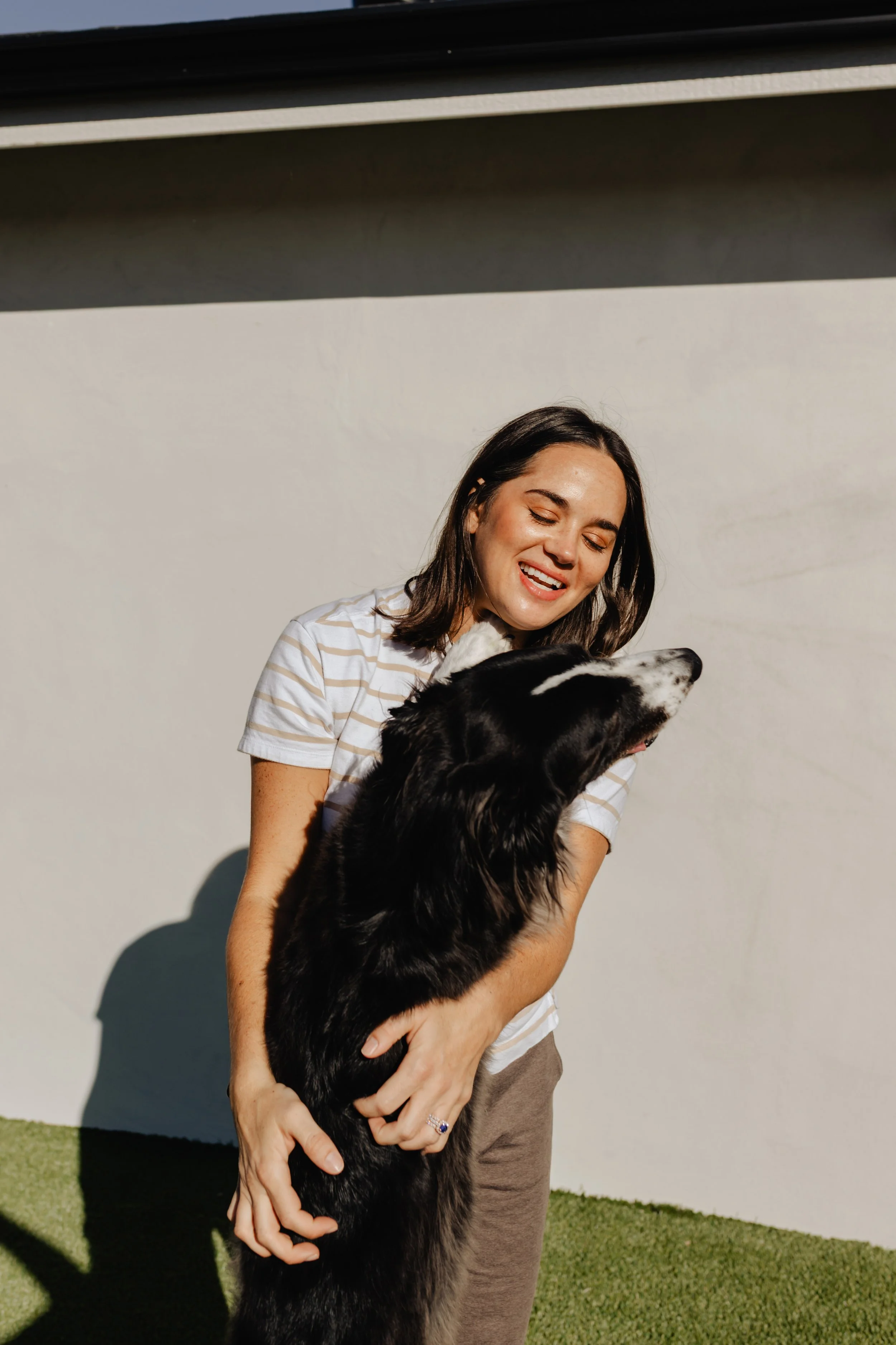 Andrea hugging a black and white border collie dog outside near a white wall, smiling.