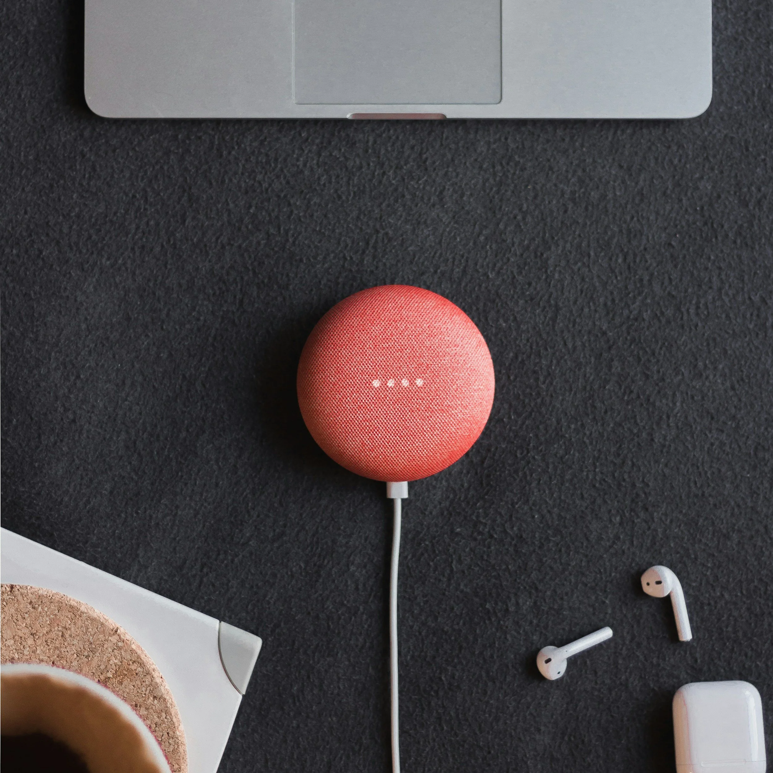 A pink portable speaker connected to a laptop on a dark surface, with wireless earbuds and a white charging case nearby.