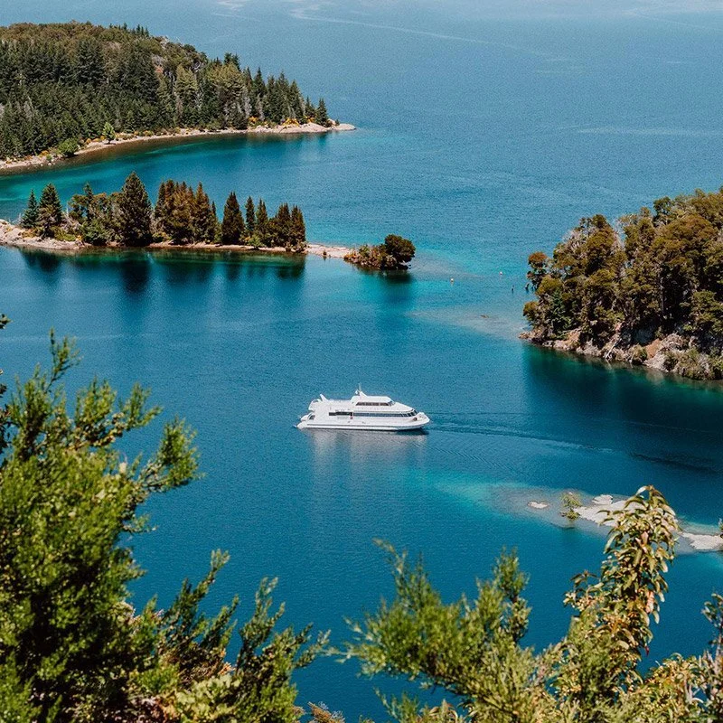 A white yacht sailing through a striking blue lake surrounded by green trees and forested hills.