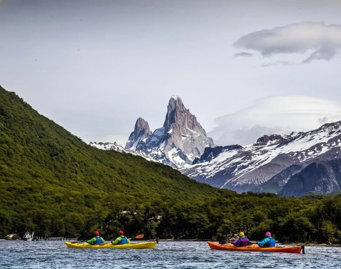 Four people kayaking on a lake with a mountain and lush green hills in the background.