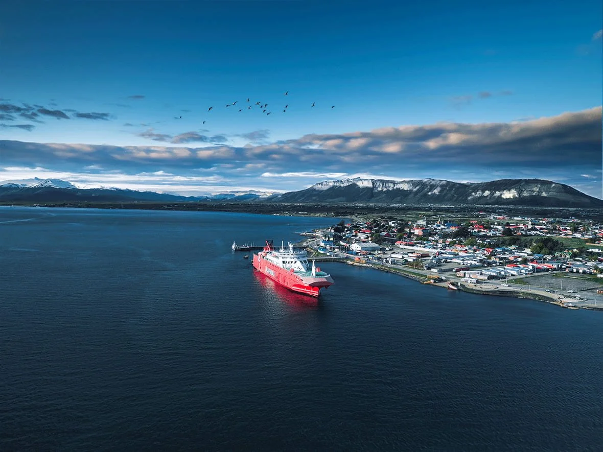Aerial view of a harbor with a large red ship docked, a coastal town with numerous buildings, and mountains in the background under a partly cloudy sky.