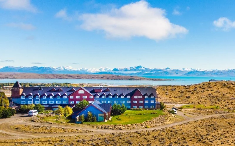 Colorful lodge with multiple stories, set in a dry landscape near a large body of water with snow-capped mountains in the background.