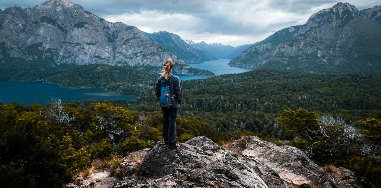 Person standing on rocky terrain overlooking a forested valley with mountains and a lake in the distance.