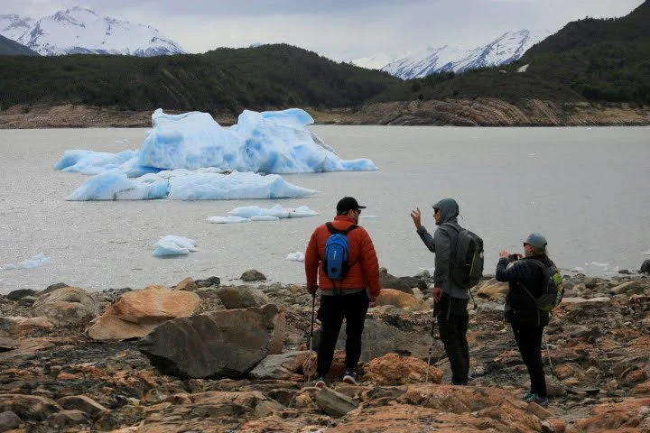 Three hikers with backpacks and trekking poles standing on rocky shore near a lake, with icebergs and snow-capped mountains in the background.