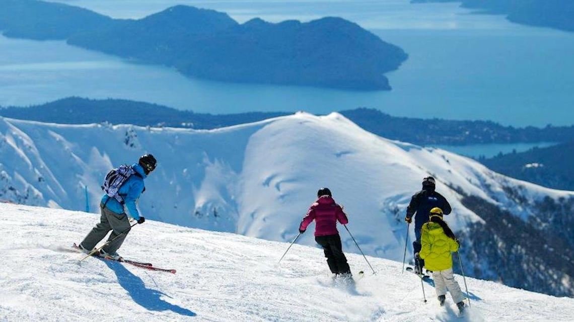 Four skiers in winter gear skiing down a snowy mountain slope with a lake and mountains in the background.
