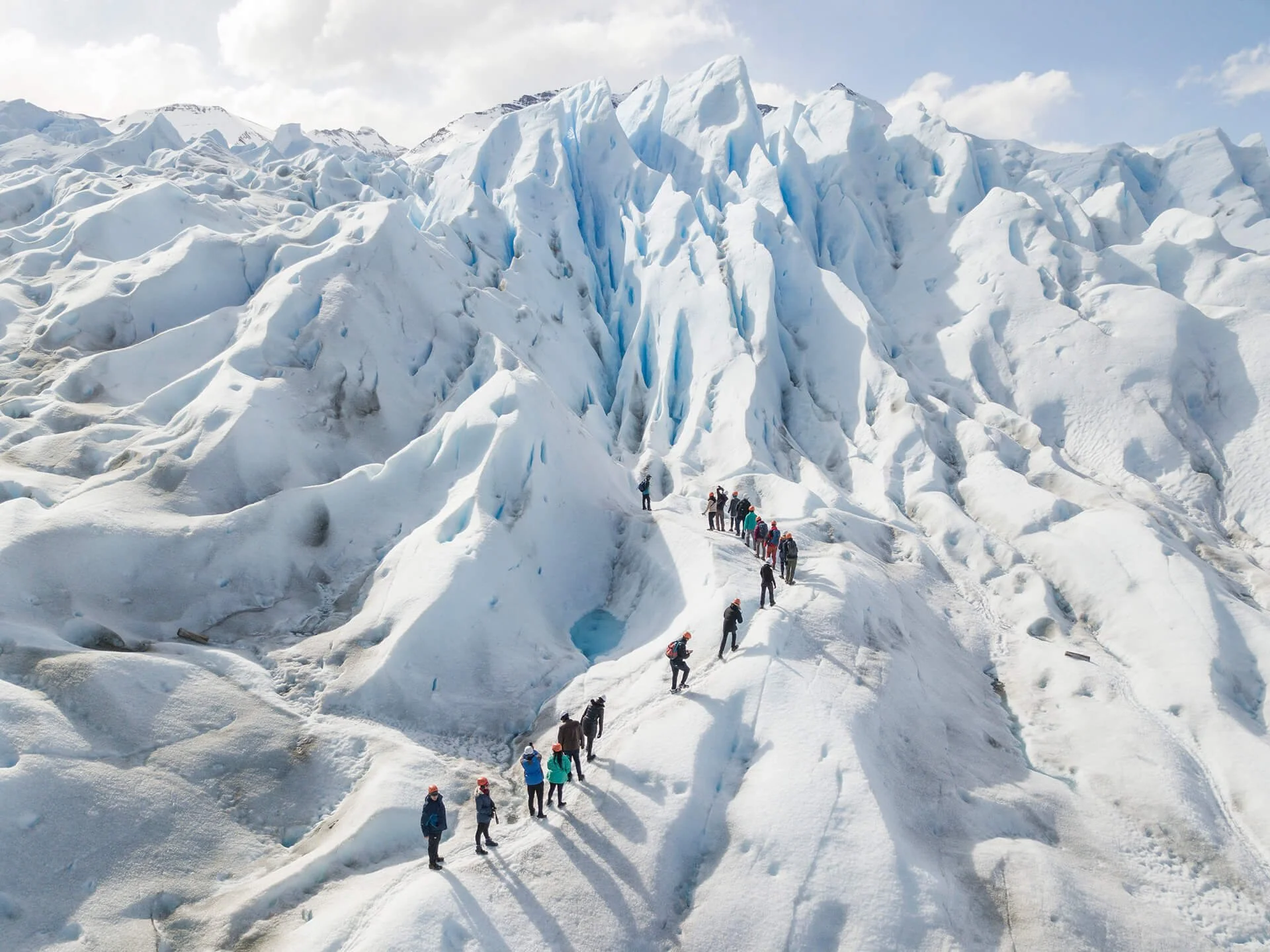 Group of hikers climbing on a glacier with large ice formations and snow-covered peaks in the background.