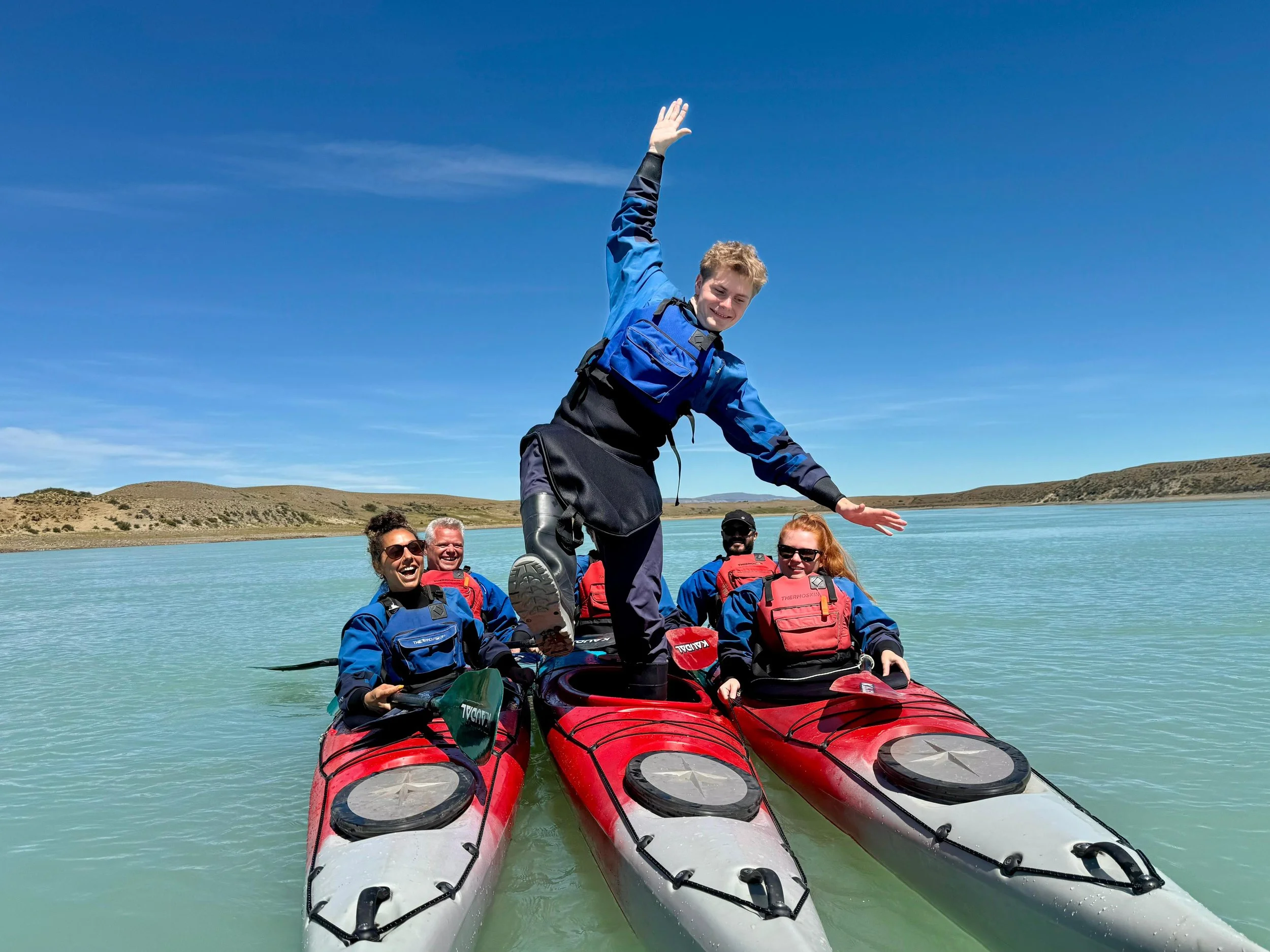 Group of five people kayaking on a calm lake, with one person standing on the kayaks, balancing and smiling amid a scenic landscape with hills and a clear blue sky.