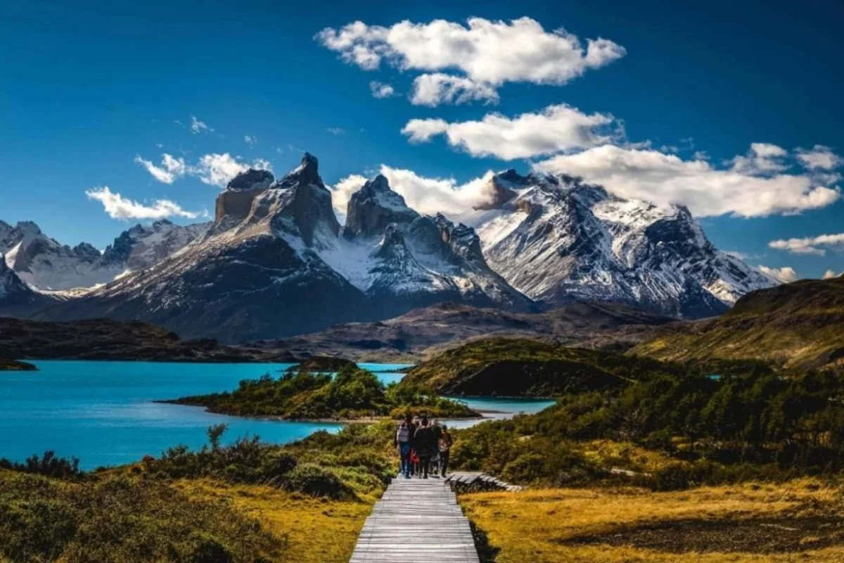A group of people walking on a wooden bridge towards a lake with turquoise water, surrounded by green bushes and mountains with snow-capped peaks under a partly cloudy sky.