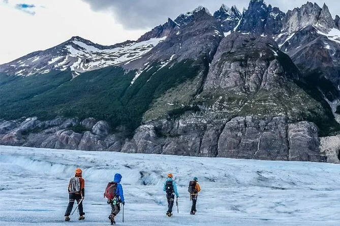 Four hikers in outdoor gear walking on icy terrain with scenic mountains and cloudy sky in the background.