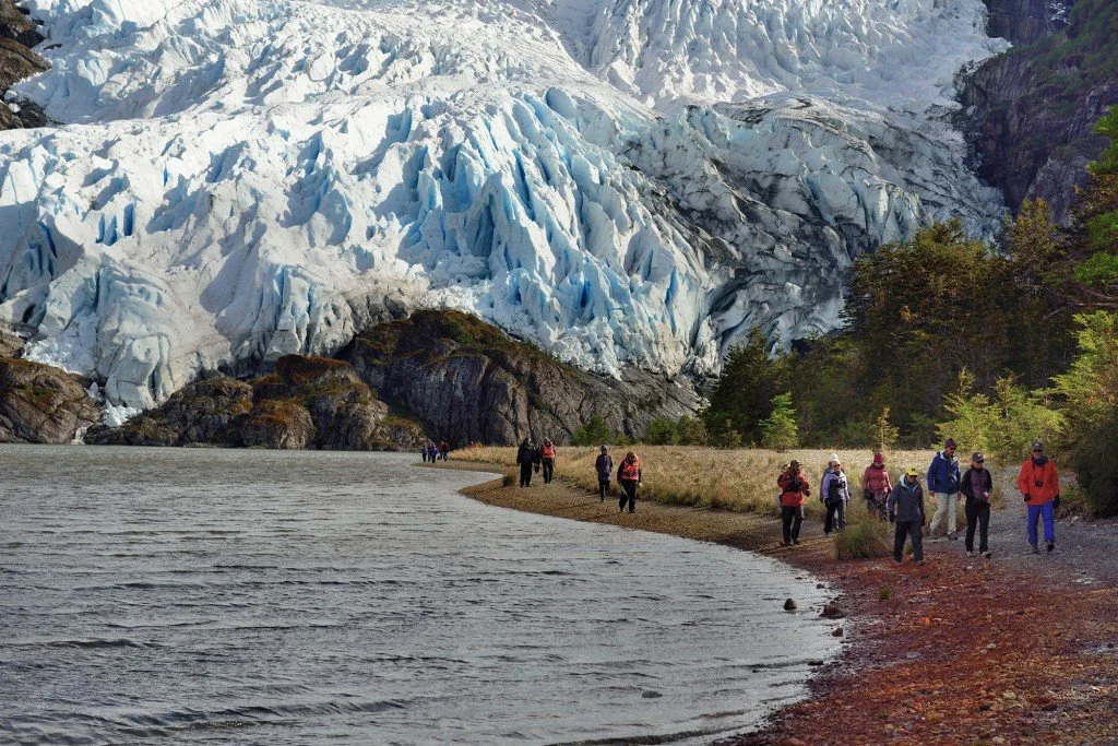 Group of hikers walking along a lakeshore with a glacier in the background.