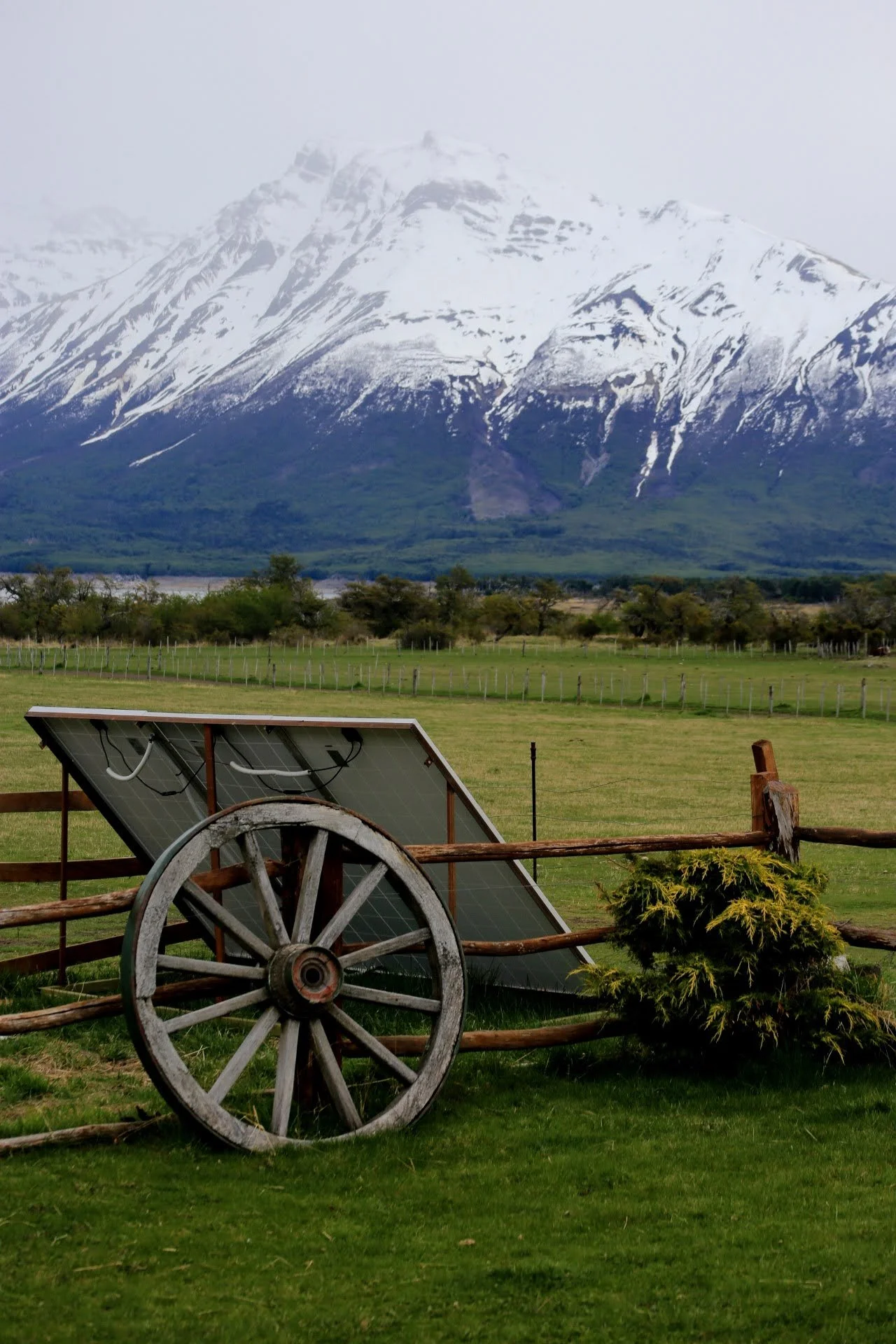 A rustic wooden wagon wheel next to a handle of a solar panel, in a green pasture with trees, and snow-capped mountains in the background.