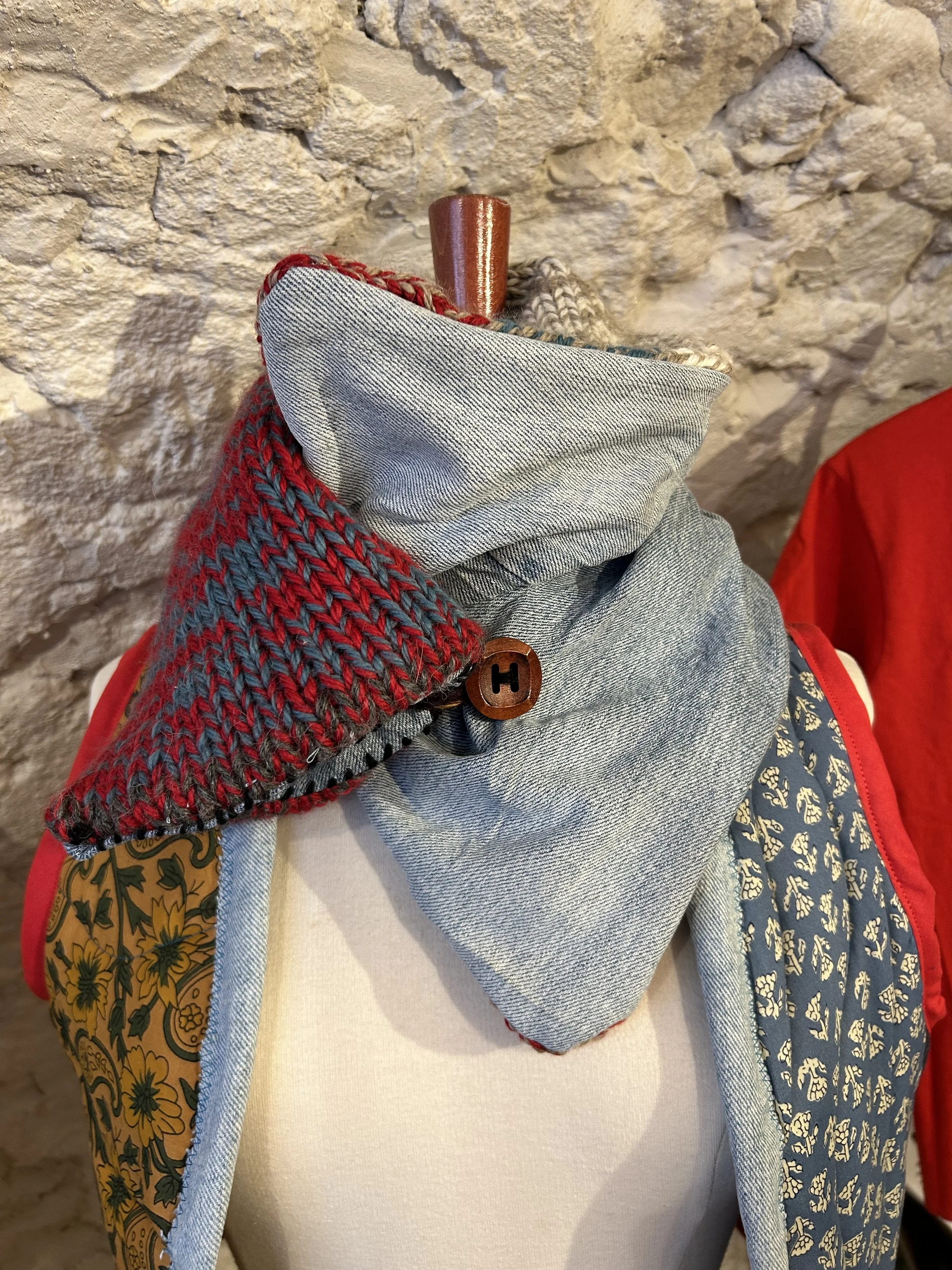 Écharpe en tissu gris avec une broche en forme de bouton marqué d'un 'H' et une pièce en laine tricotée rouge et grise, exposée sur un mannequin avec un mur en pierre en arrière-plan.