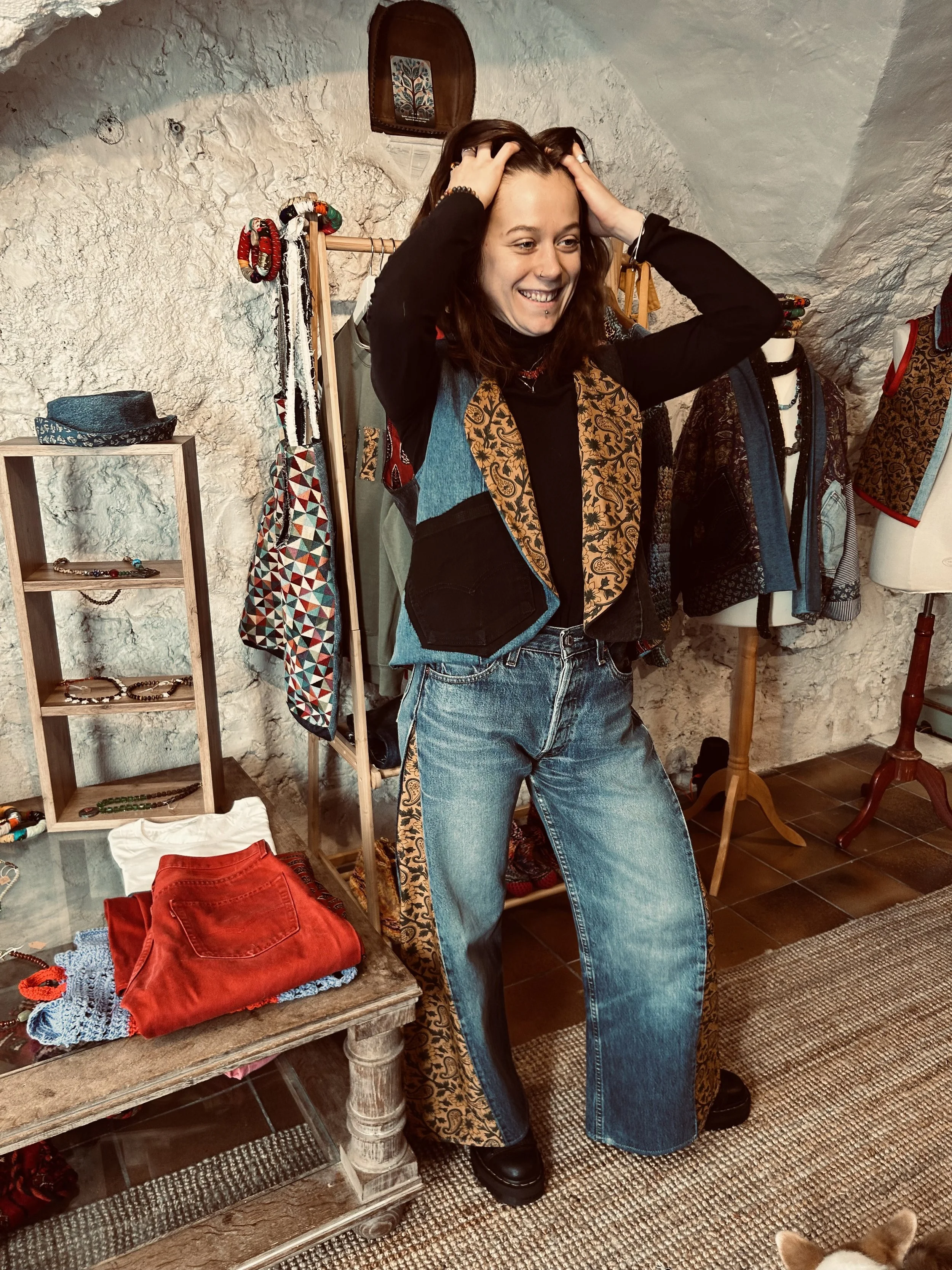 Jeune femme souriante posant dans une boutique de vêtements vintage, avec des vêtements et accessoires exposés autour d'elle.