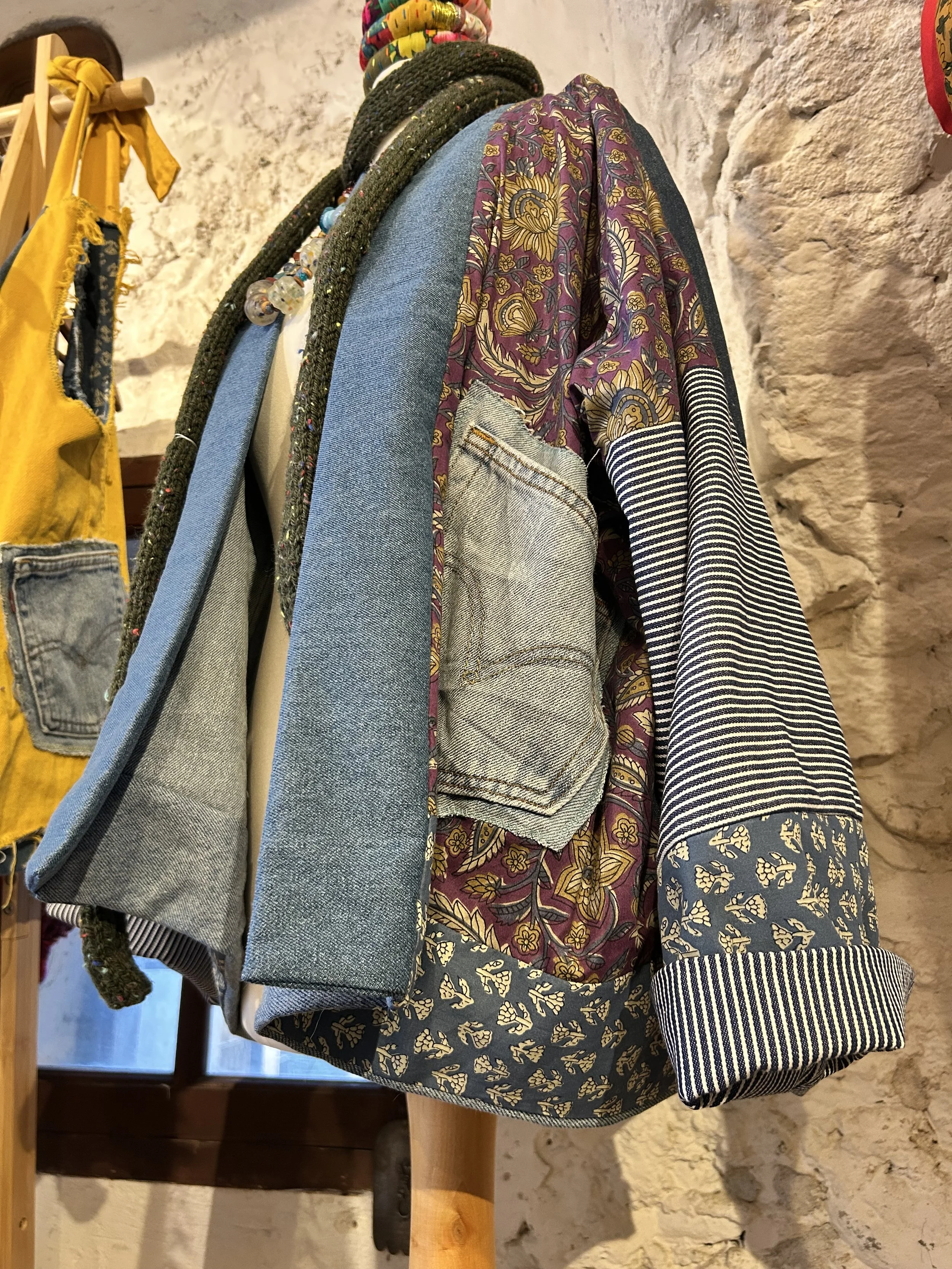 Vêtements suspendus comprenant une veste en jean, un blazer beige, un chemisier avec motif floral, une veste à rayures, un pantalon avec motif floral, et divers accessoires comme un collier, contre un mur en pierre.