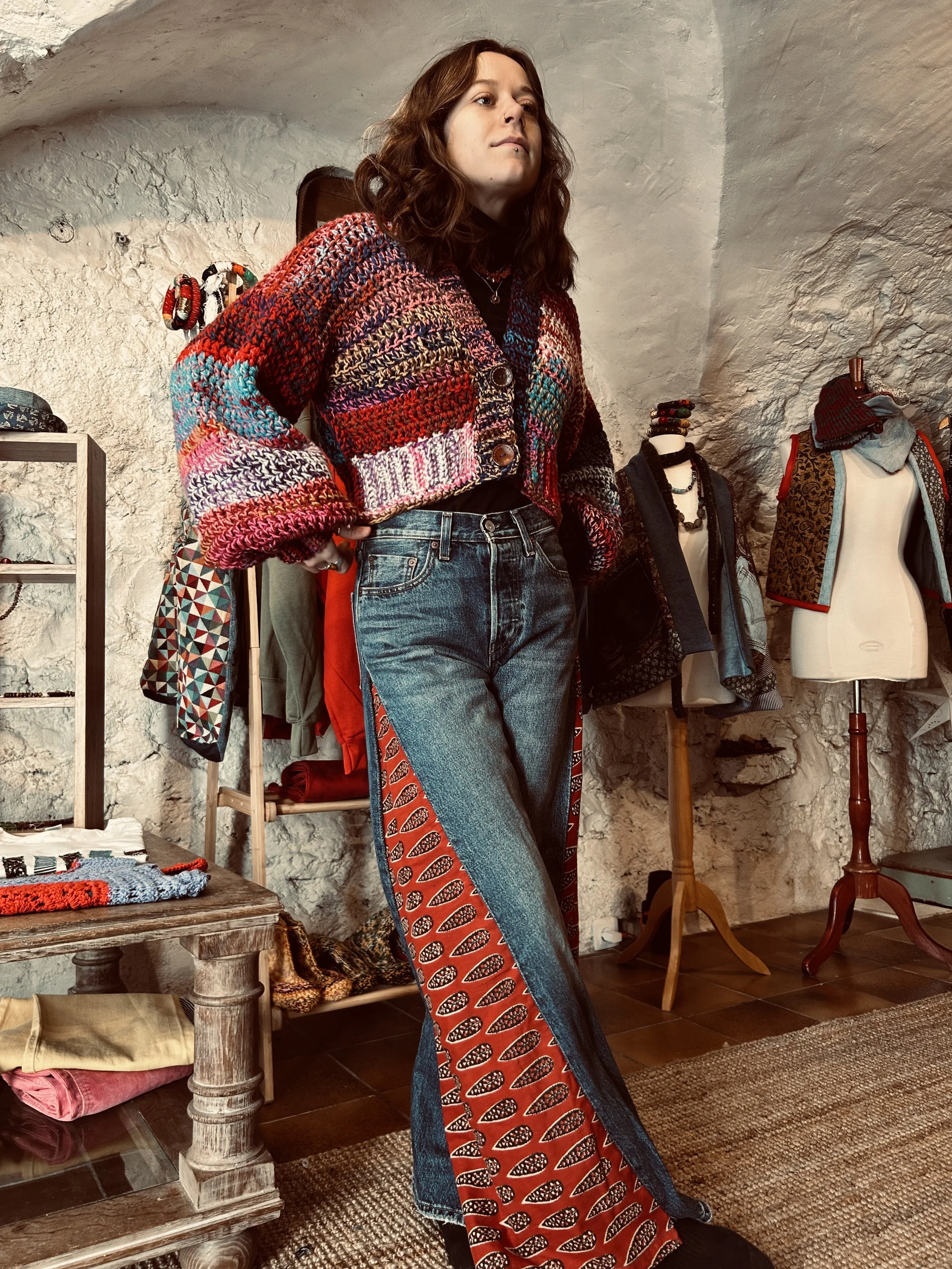 Une femme debout dans une boutique de vêtements, portant un cardigan multicolore et un jean avec un motif rouge et noir sur le côté. Autour d'elle, des mannequins et des vêtements exposés.
