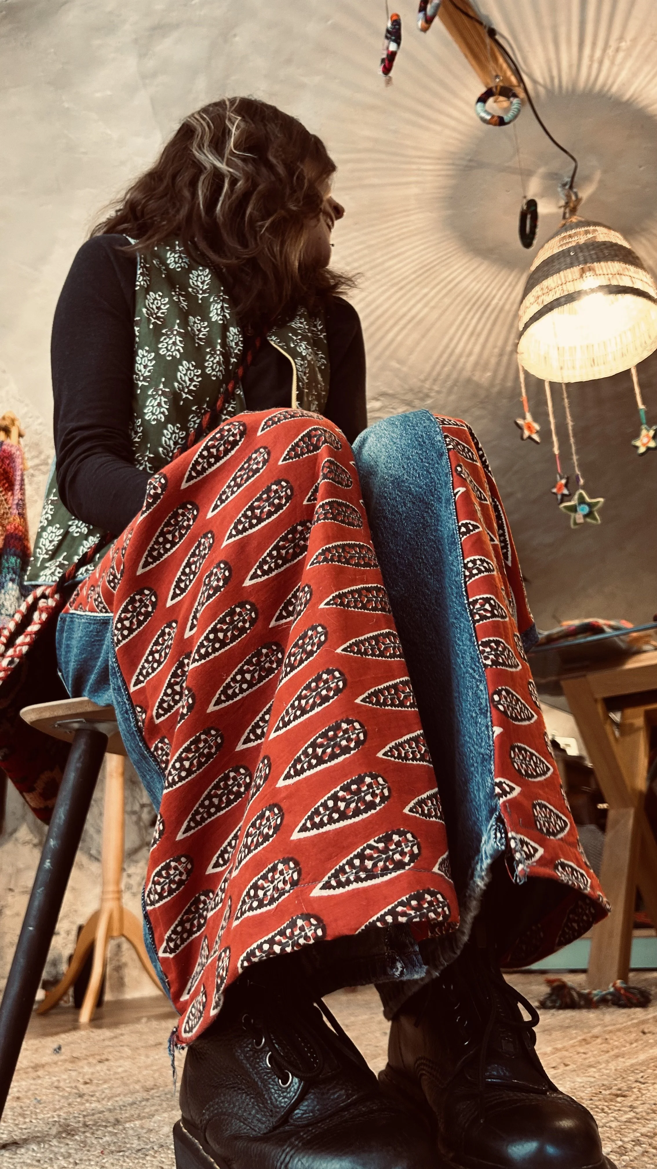 Une femme assise sur une chaise, vue de bas, portant un pantalon rouge avec un motif noir et blanc, des bottes noires, dans une pièce avec un plafond en pierre et une lampe suspendue.