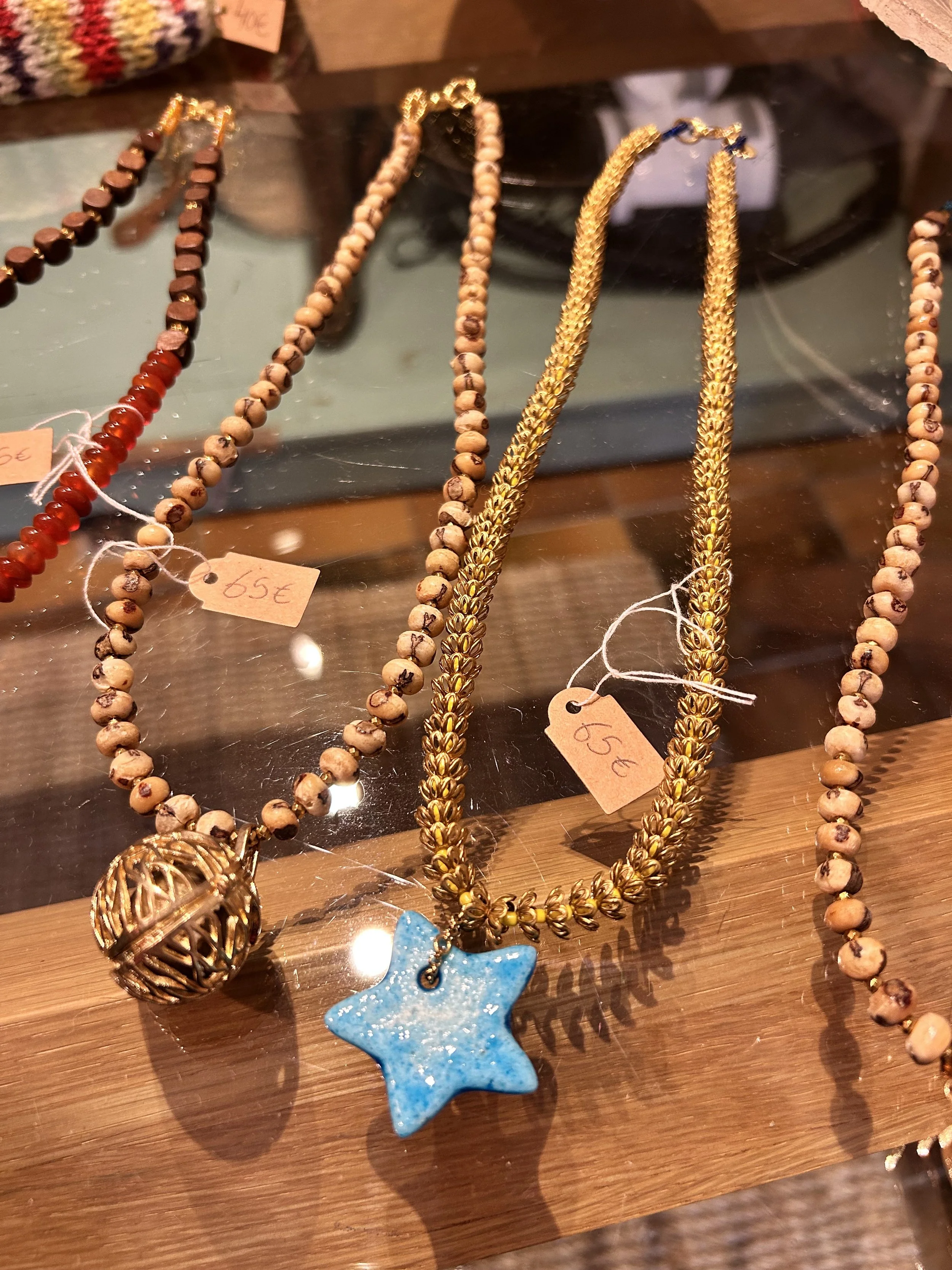 Colliers élégants avec bijoux fantaisie, dont un collier avec un pendentif étoile en céramique bleue et un autre avec une boule en métal doré, exposés sur un support en bois dans une vitrine.