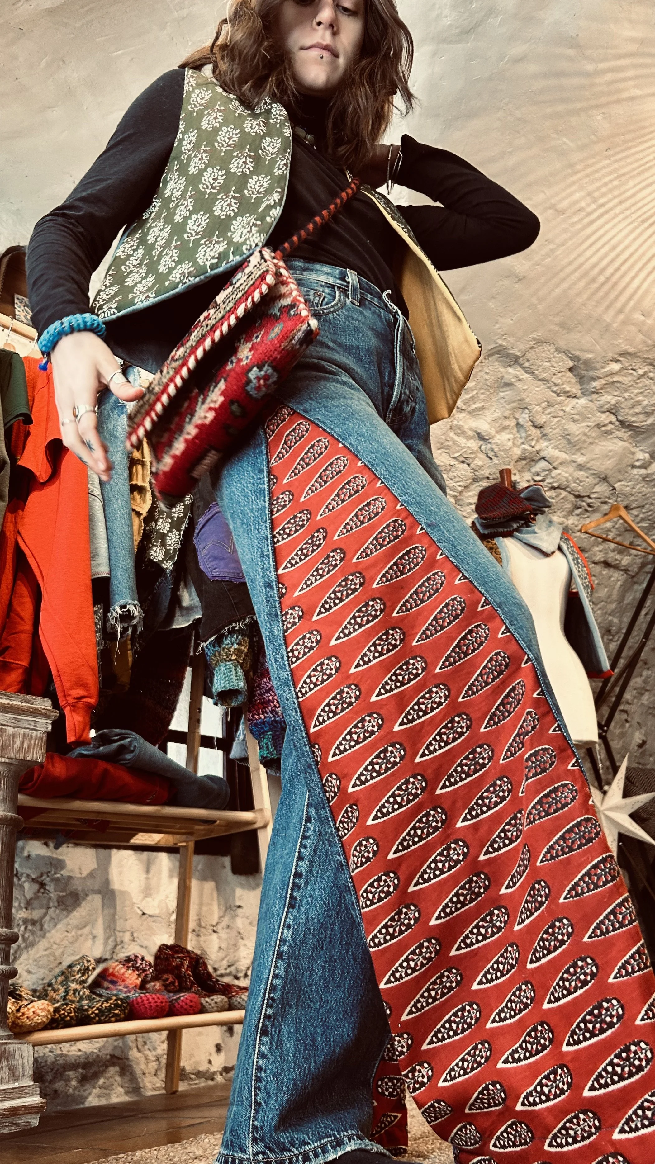 Une femme porte un pantalon en jeans avec un motif rouge et noir, un haut noir, et une veste ton sur ton. Elle possède des cheveux bruns bouclés et porte un piercing au labret. La scène semble être dans un magasin de vêtements vintage ou de seconde main, avec diverses fringues exposées en arrière-plan.