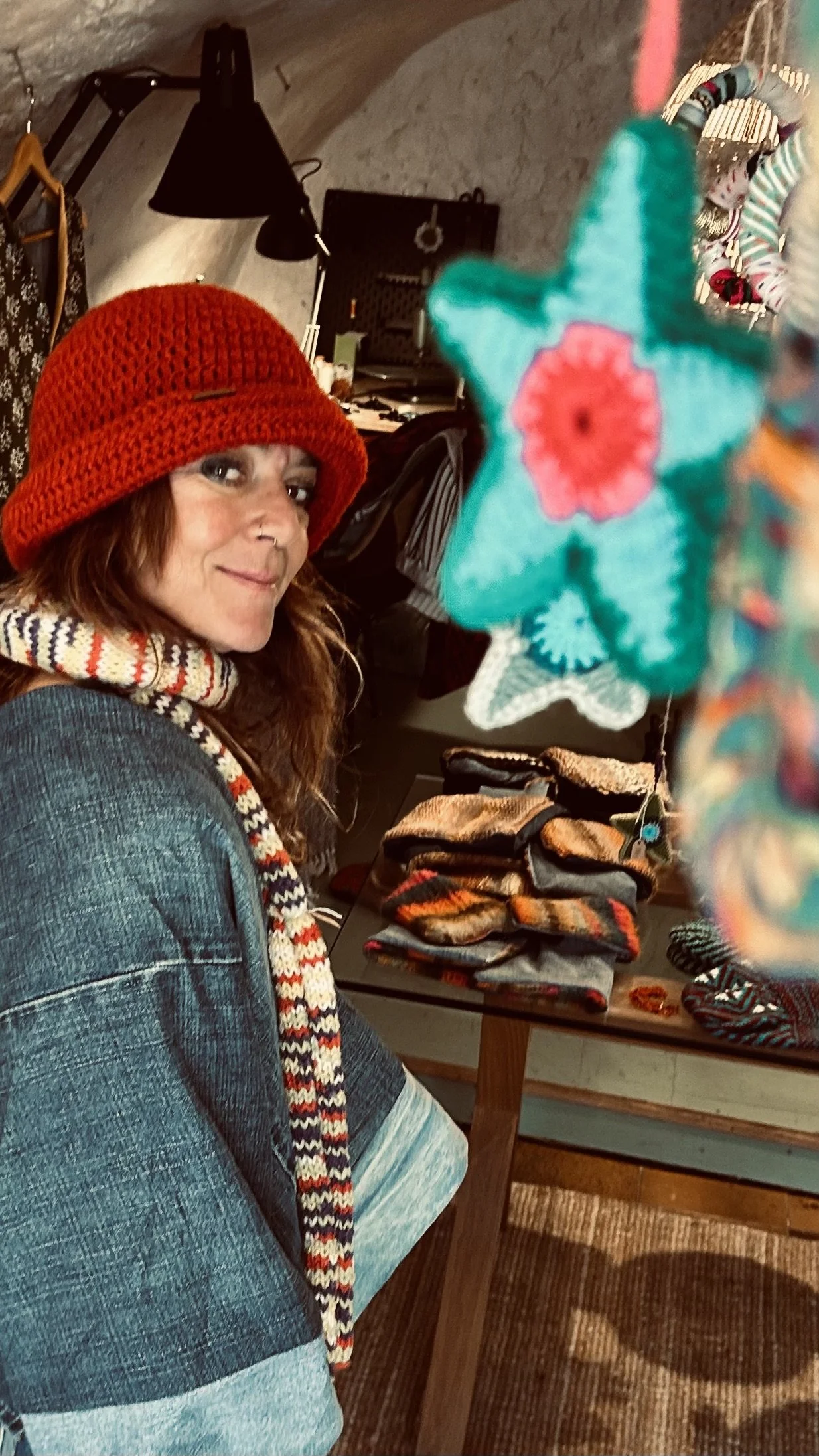 Femme portant un chapeau rouge, debout à côté d'un étalage d'accessoires comme des gants et des couvre-chefs, dans un intérieur avec un plafond en pierre, décoré avec des objets artisanaux et textiles colorés.