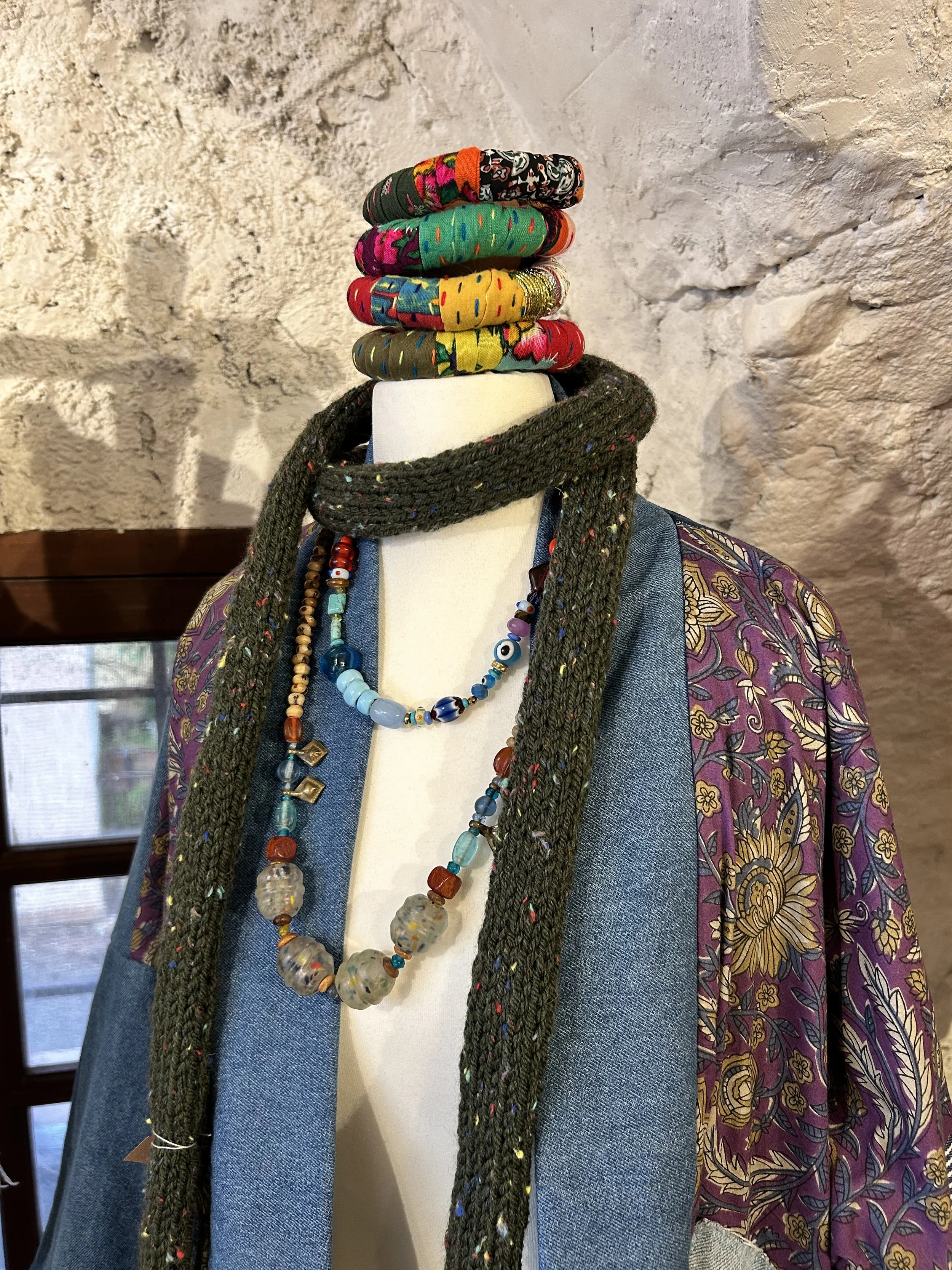 Mannequin portant plusieurs bracelets colorés, un collier en perles, un foulard gris, une veste en denim et une blouse à motifs floraux violets et jaunes, avec un mur en pierre en arrière-plan.