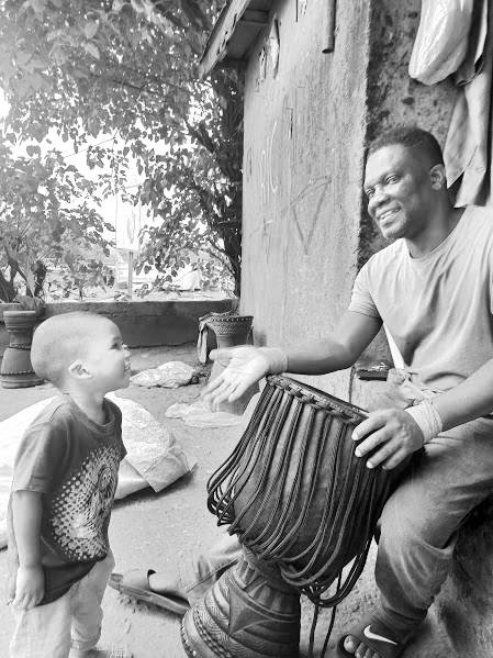 A man sitting outdoors extends his hand to a young boy with short hair, wearing a T-shirt, as they share a moment of greeting. The man is holding a traditional musical instrument, possibly a balafon, and there are plants and trees in the background.