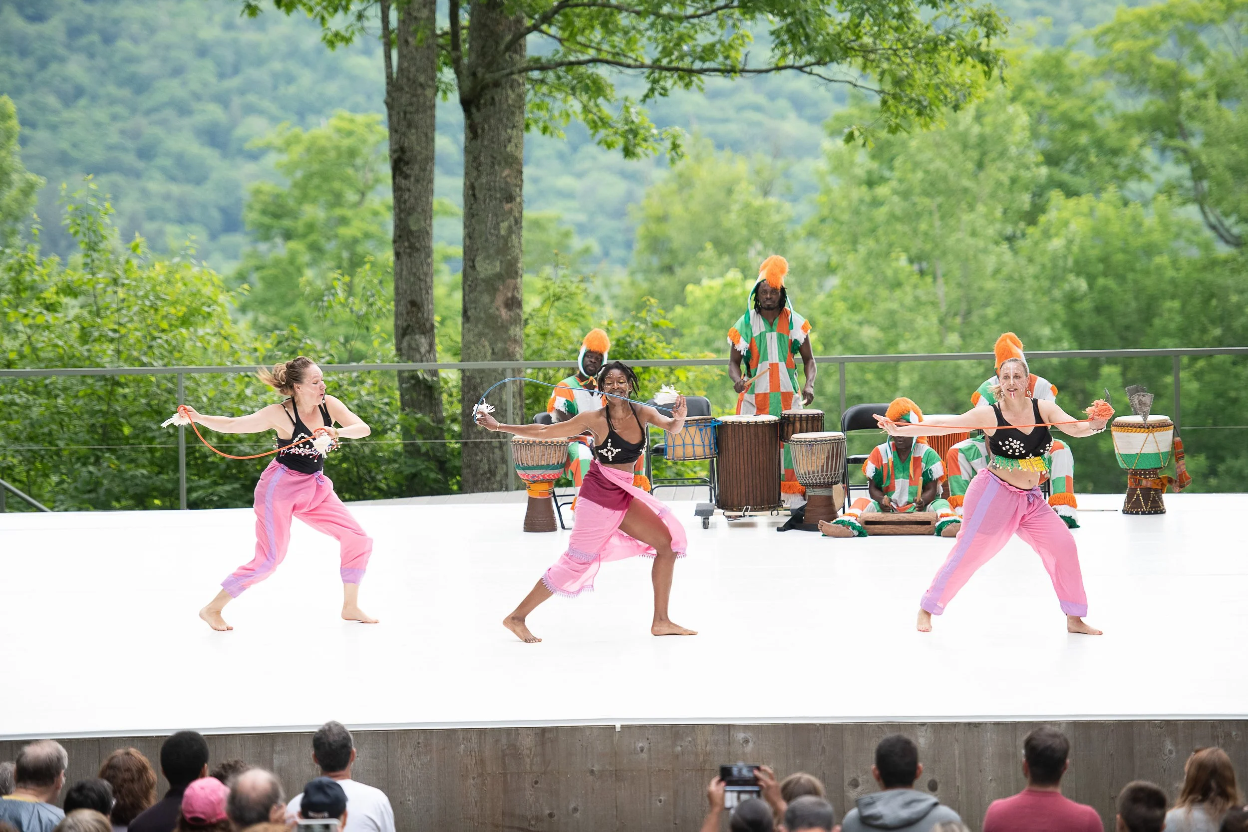 Dancers performing a traditional dance on stage with musicians playing drums in the background, outdoors with green trees.