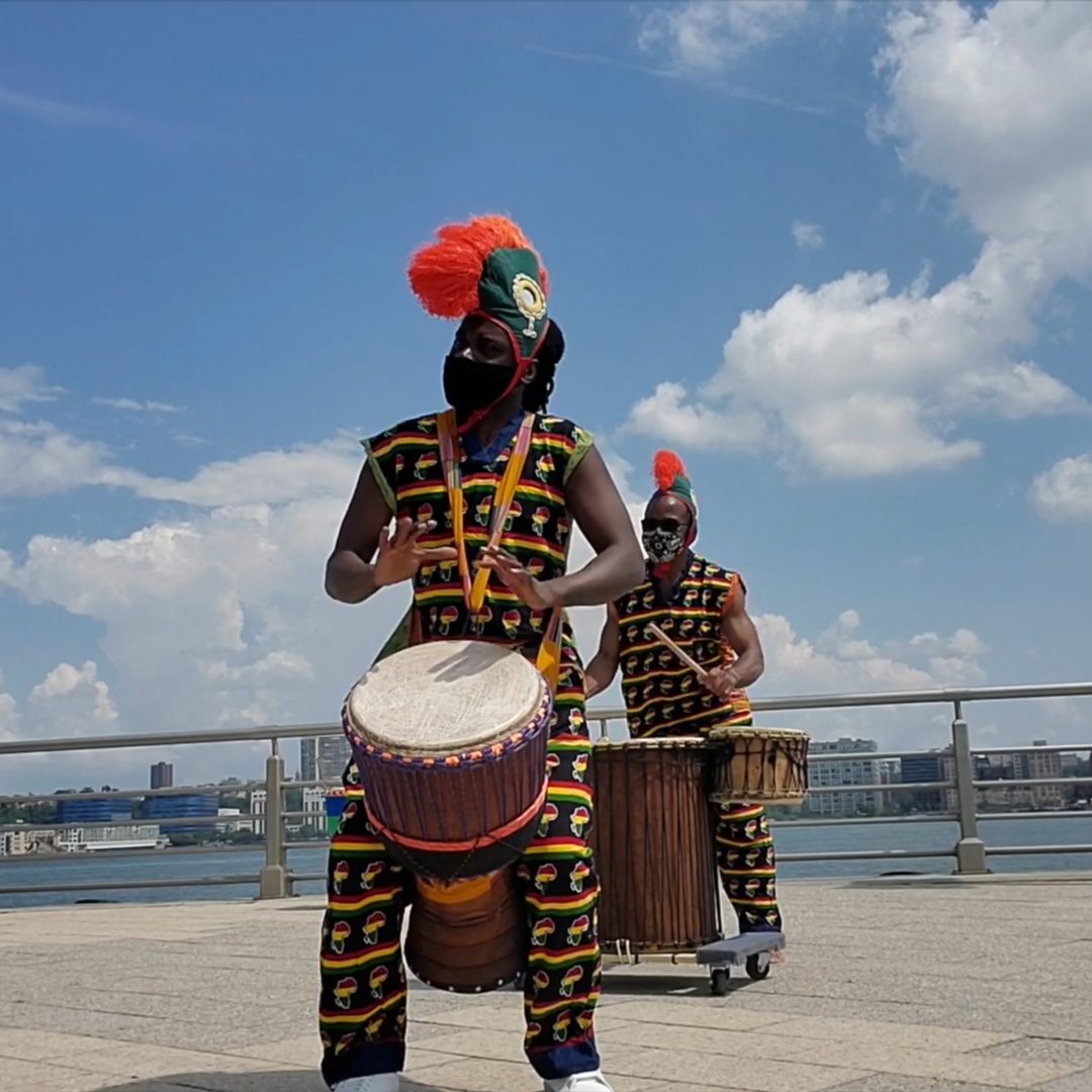 Two performers dressed in colorful traditional African attire with feathered headdresses, playing drums outdoors against a cityscape and cloudy sky.