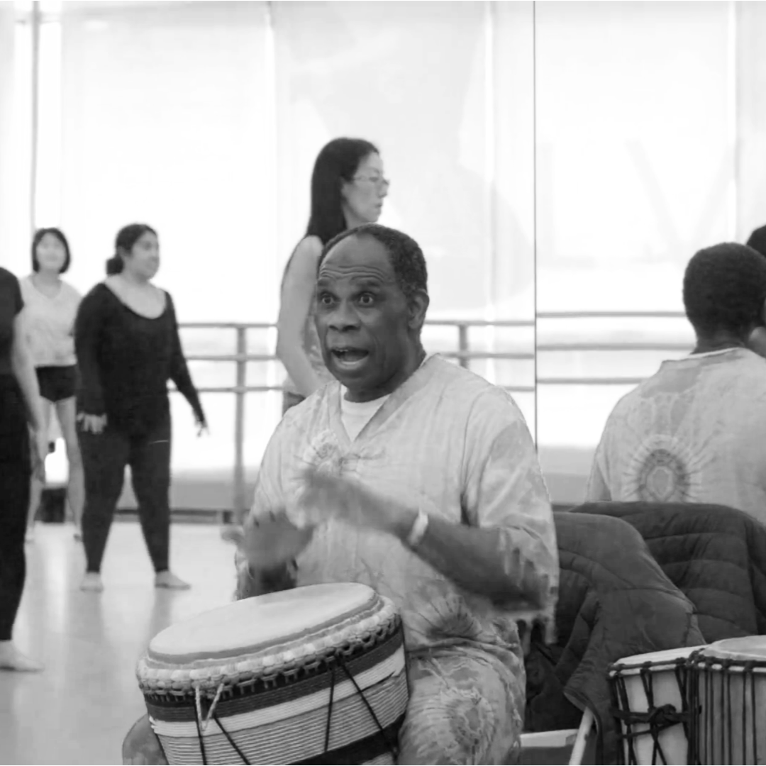 A Black man playing the djembe drum in a dance studio, with a woman and other people in the background.