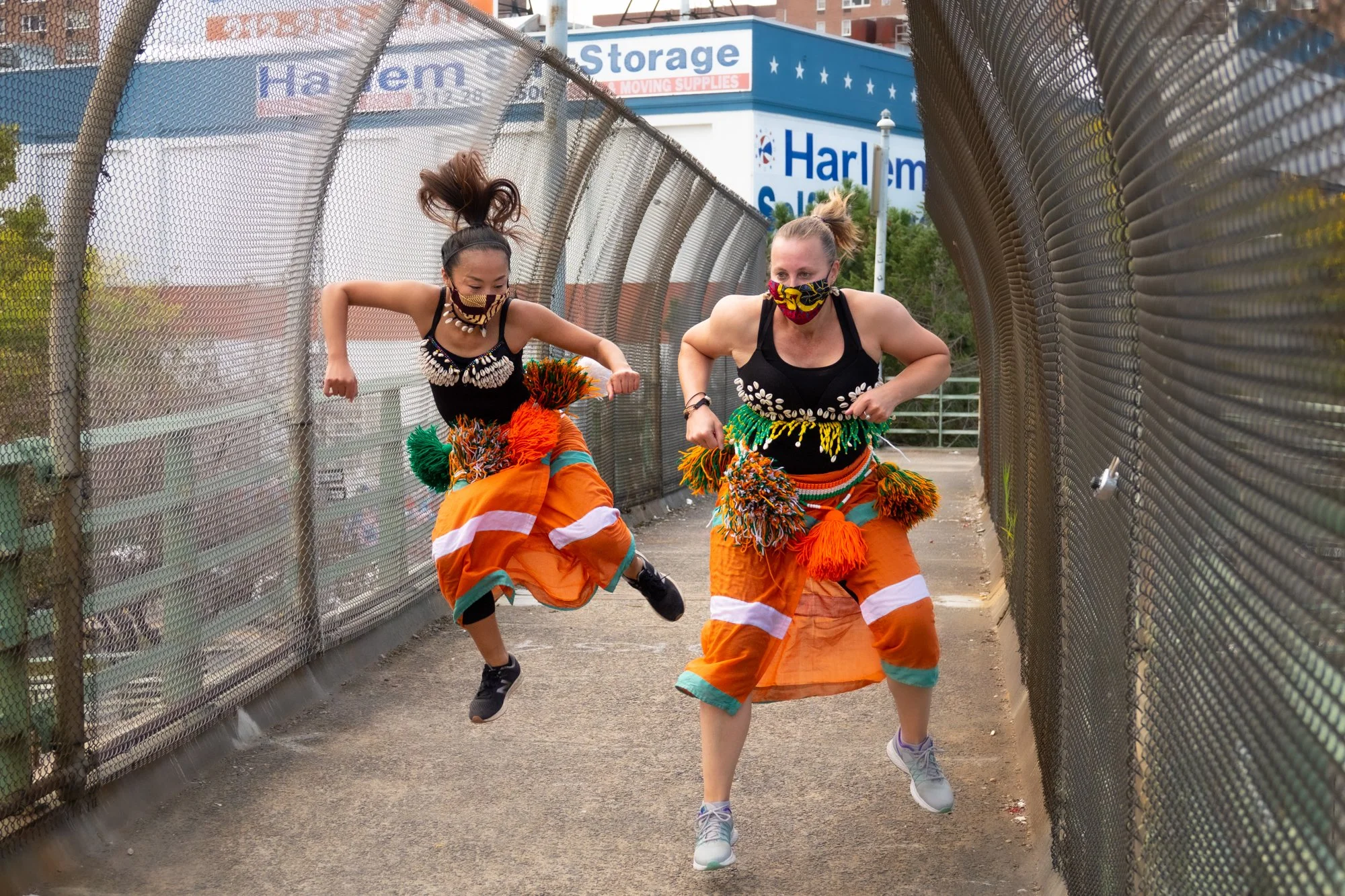 Two women in colorful, festival-style clothing and face masks jumping in the air along a pedestrian bridge.