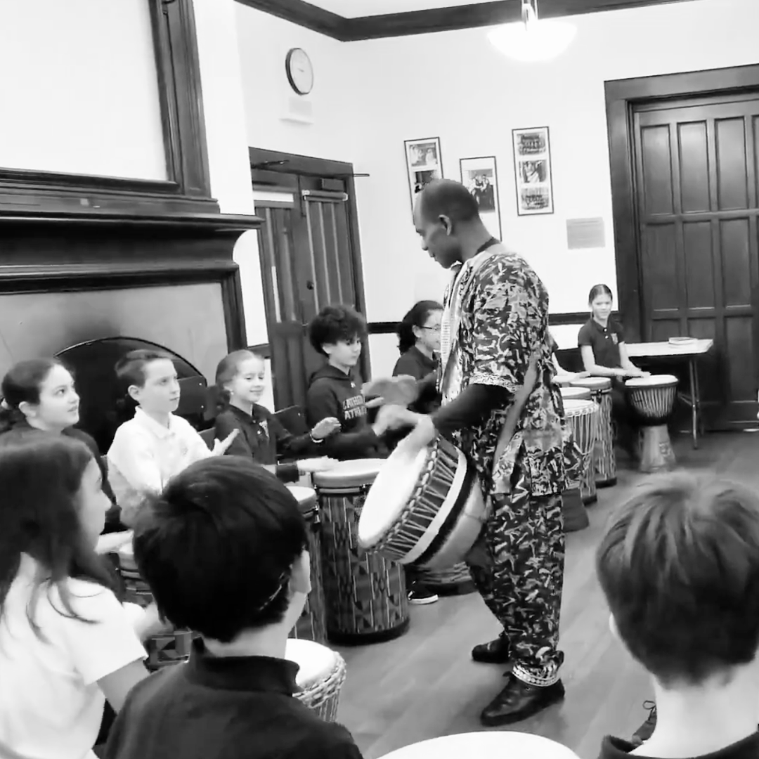 A person in patterned clothing is playing a drum in front of a group of children seated in a semi-circle, each with a drum or percussion instrument in a classroom or community room.