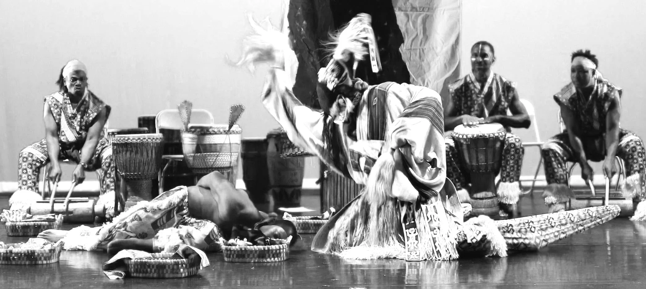 A traditional African drum performance featuring five men dressed in patterned attire. One man is in a dynamic dance pose in front of a seated audience. Drum baskets, drums, and musical instruments are on stage.
