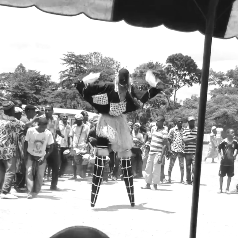 A performer in a traditional costume on stilts entertains a crowd at an outdoor event, with trees in the background.