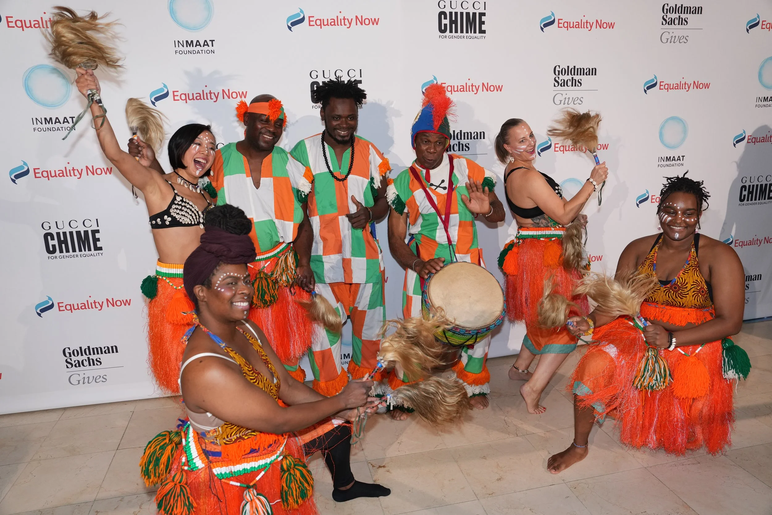 Group of eight people dressed in colorful traditional African attire, dancing and smiling at an indoor event with a backdrop displaying logos of INMAAT Foundation, Equality Now, Gucci Chime, and Goldman Sachs.