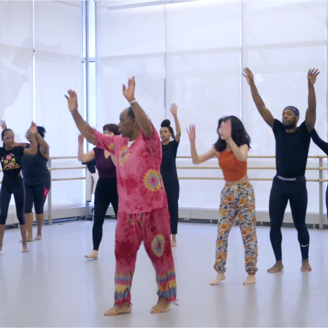 Group of diverse people in a dance studio participating in a dance class, following instructor's movements, with hands raised.